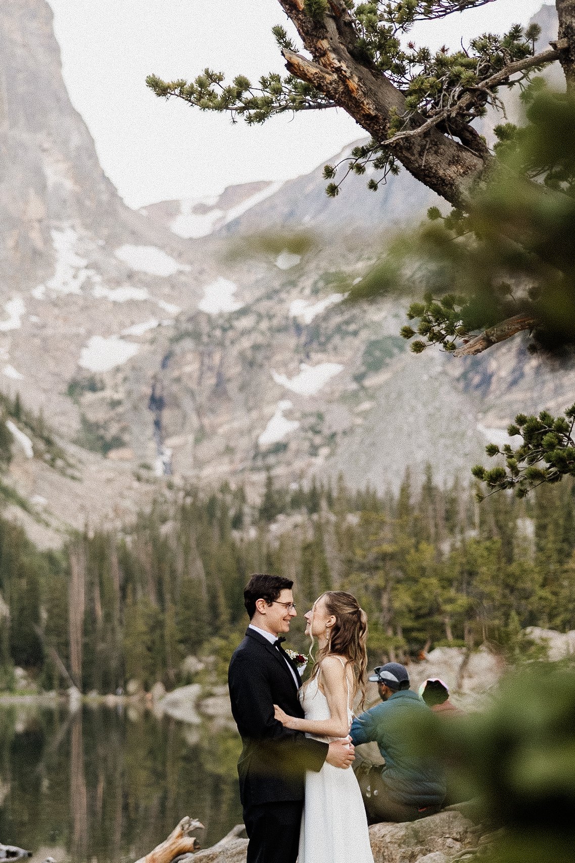 Unedited photo of a couple embracing by a mountain lake, with a fisherman visible in the background