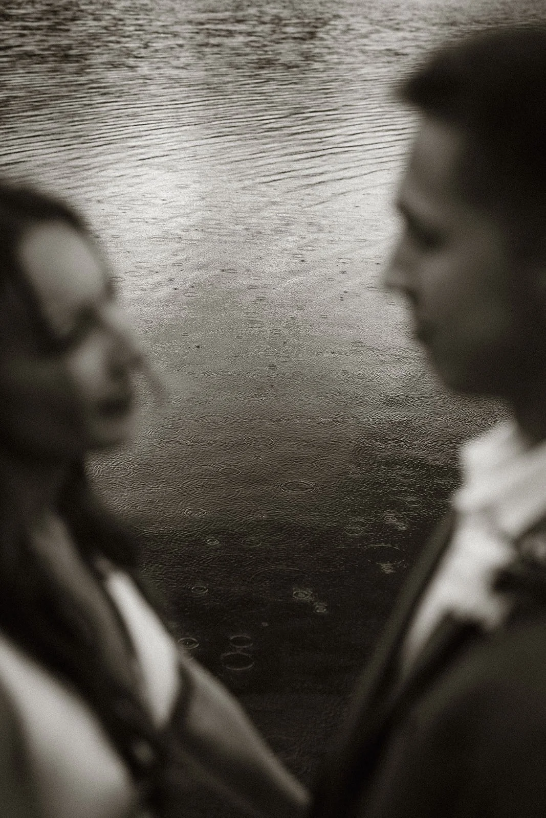 A moody black and white portrait of a couple during a rain shower, their reflections shimmering on the wet pavement. The image captures an intimate and emotional moment of connection amidst the rain, highlighting a unique wedding photo style.