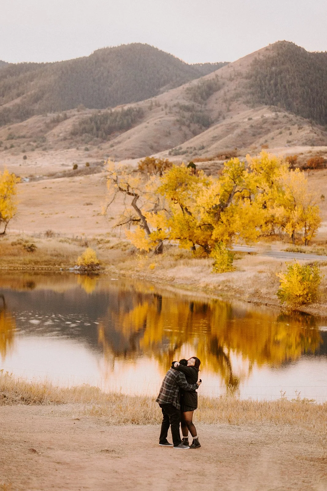 A newly engaged couple hugging at the edge of a pond reflecting golden cottonwoods