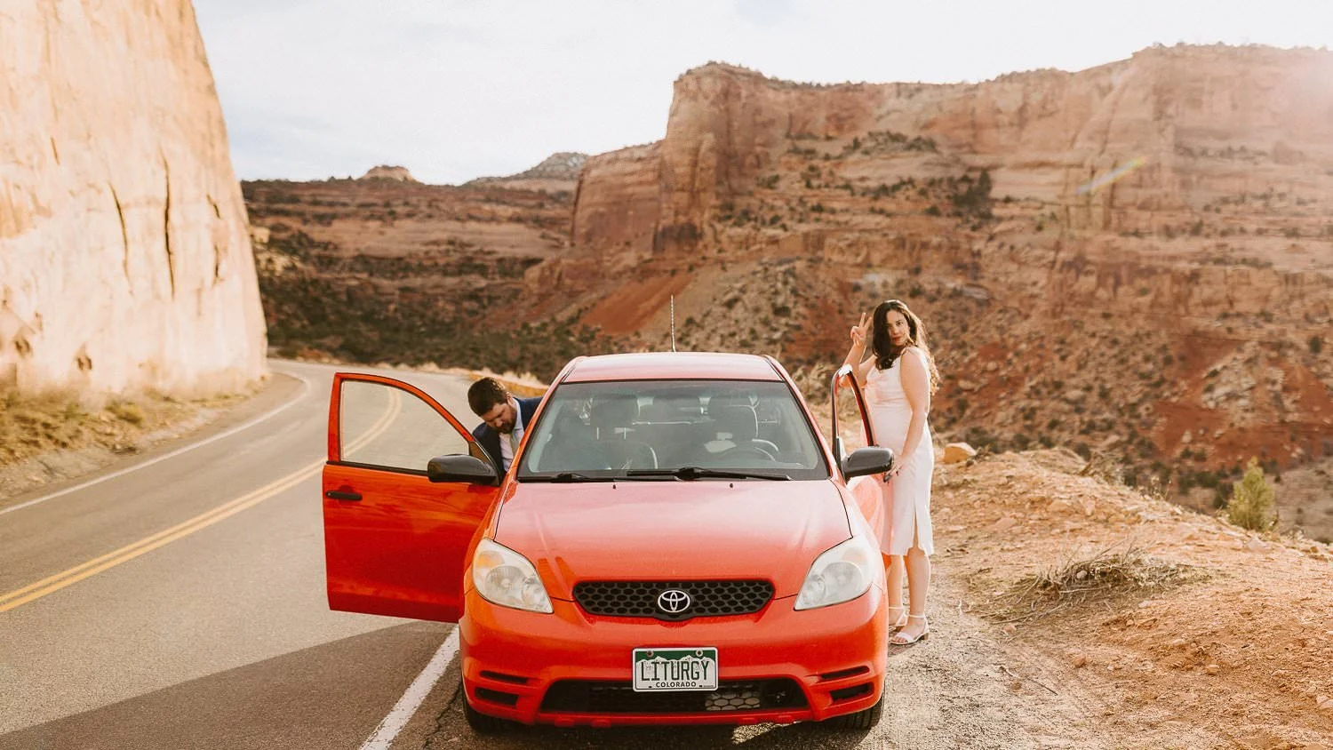 A bride and groom standing next to a red car in a desrt canyon give a peace sign