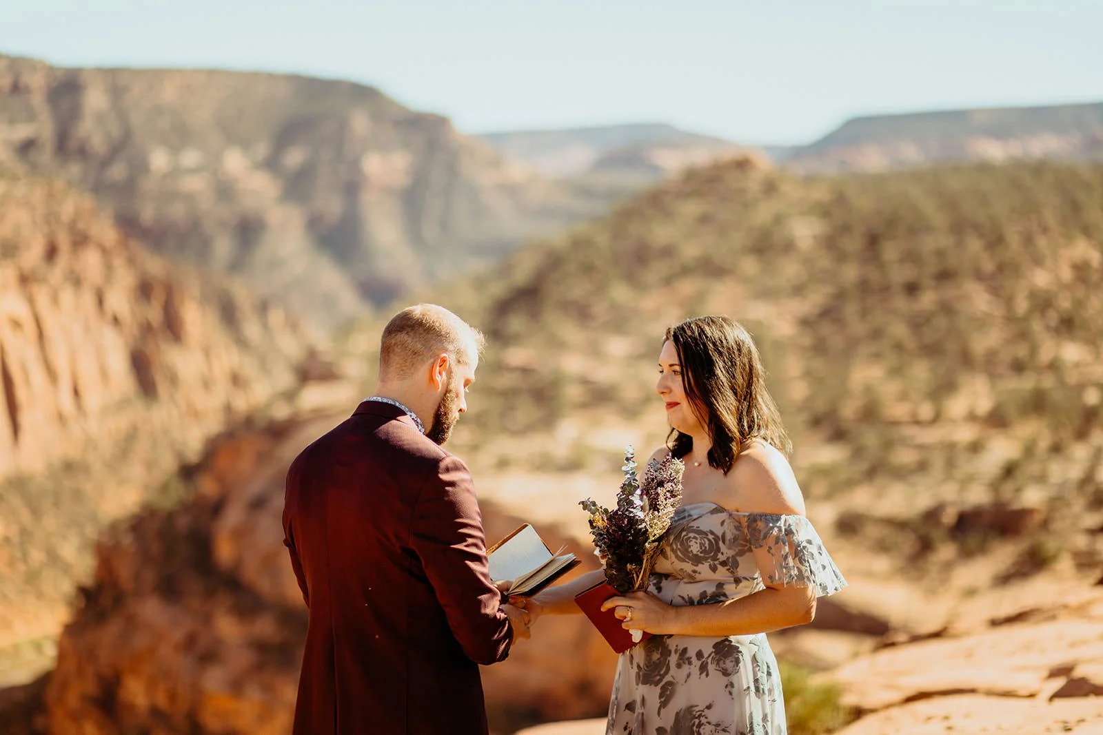 Elopement vows on a red rock canyon overlook, with a blue floral off the shoulder dress and a burgundy suit
