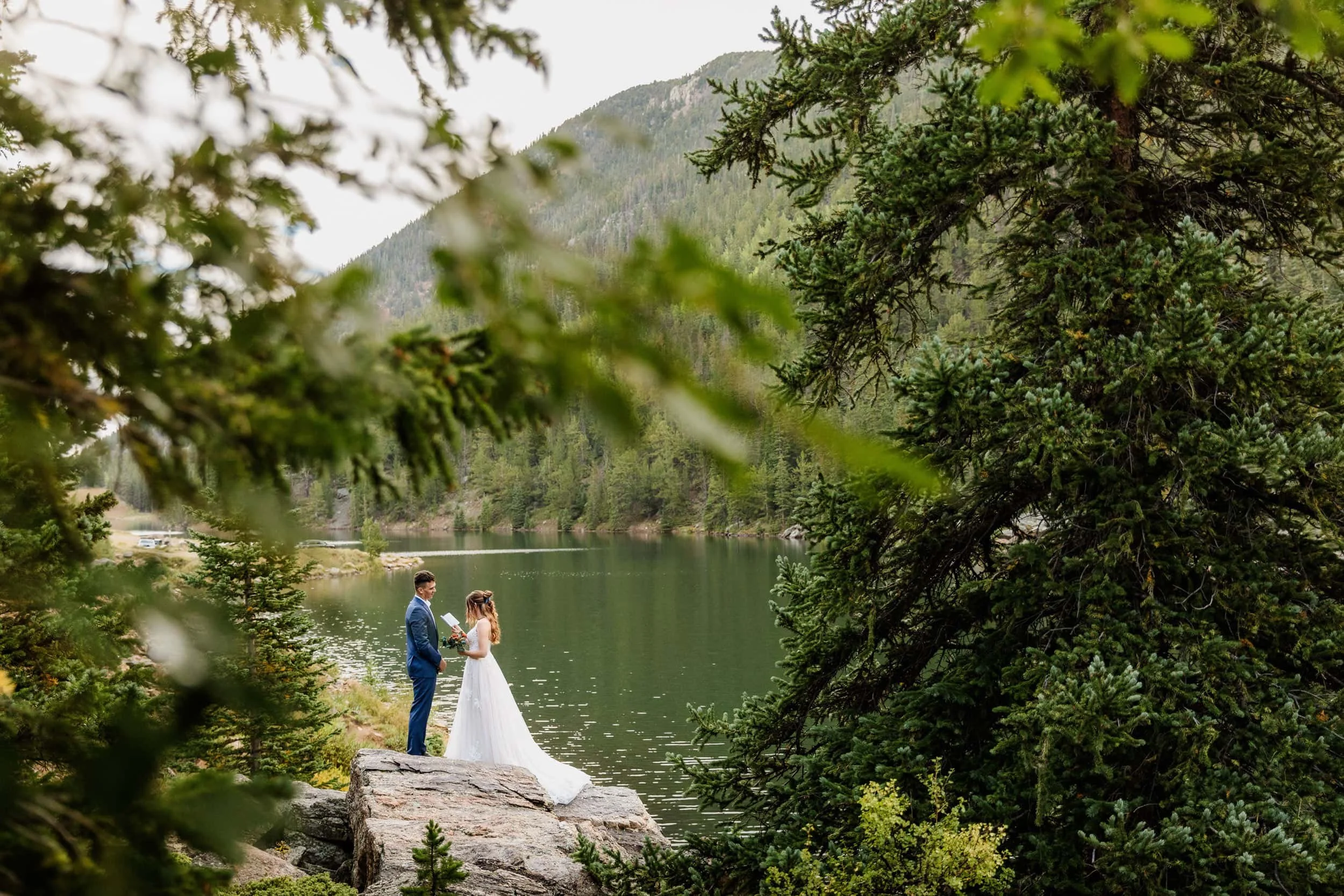 Wedding ceremony by an alpine lake surrounded by mountain peaks, framed by pine tree branches