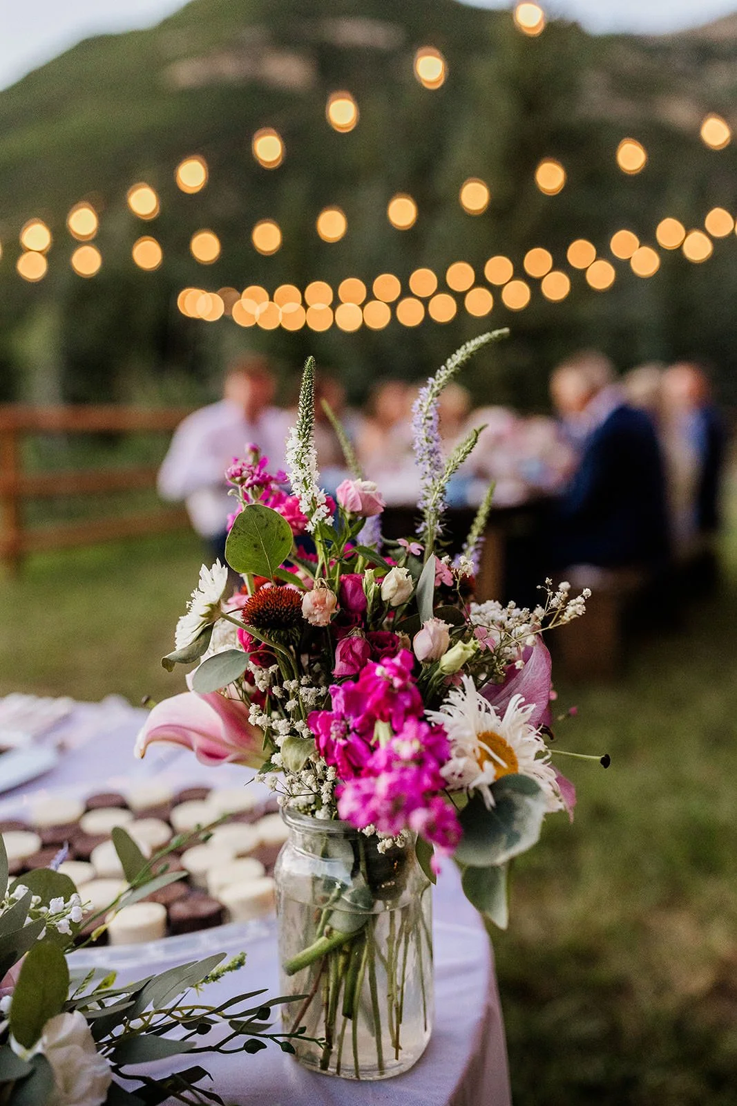 Colorful floral centerpiece in a glass jar at an outdoor gathering, with blurred string lights and people in the background.