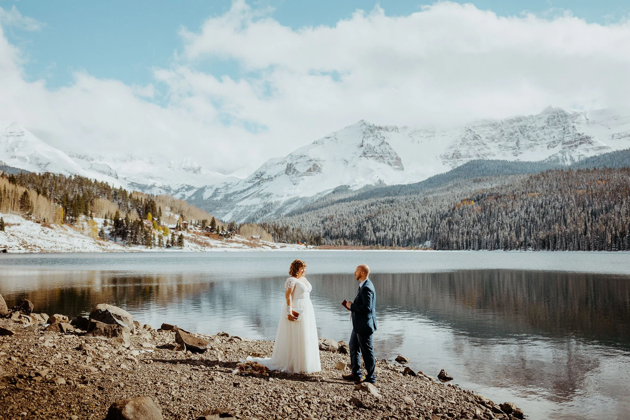 A couple faces each other on the shore of an alpine lake near Telluride with snowy peaks behind them