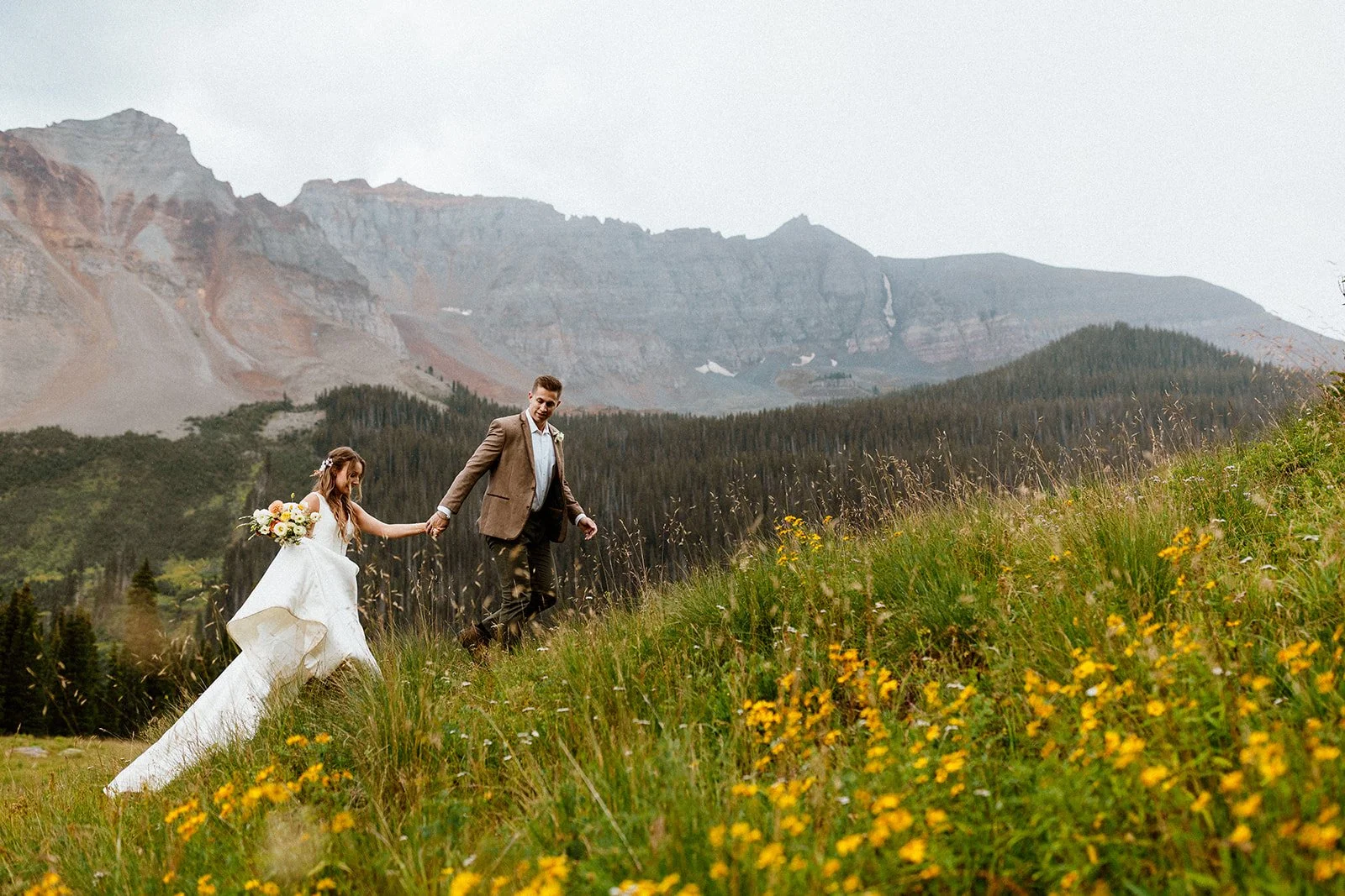 A bride and groom in wedding clothes hold hands leading each other up a hill covered in wildflowers in front of a mountain range