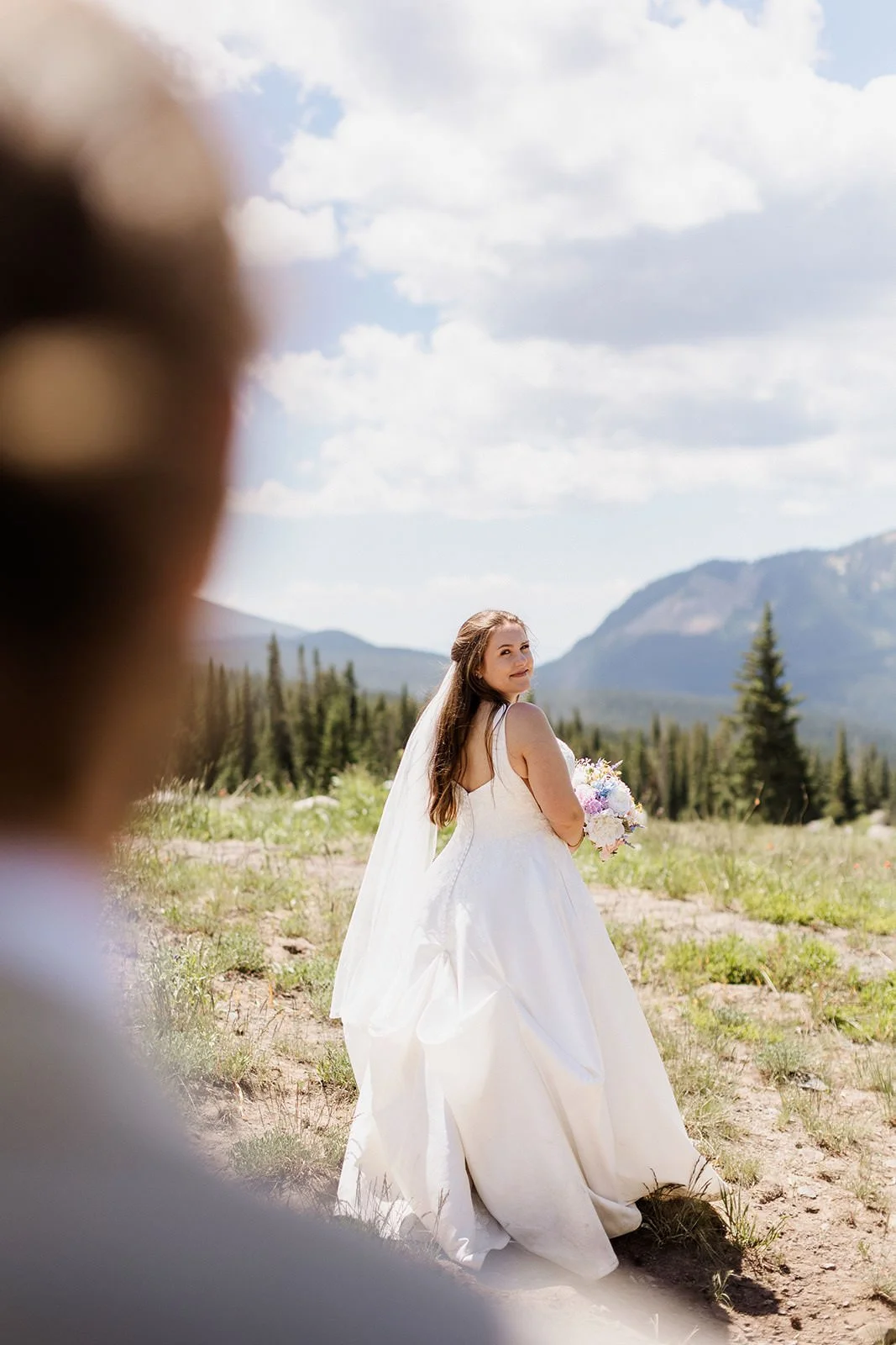 A bride in a white wedding gown holding a bouquet, standing outdoors in a mountainous landscape with evergreen trees and partly cloudy sky, smiling and looking back.
