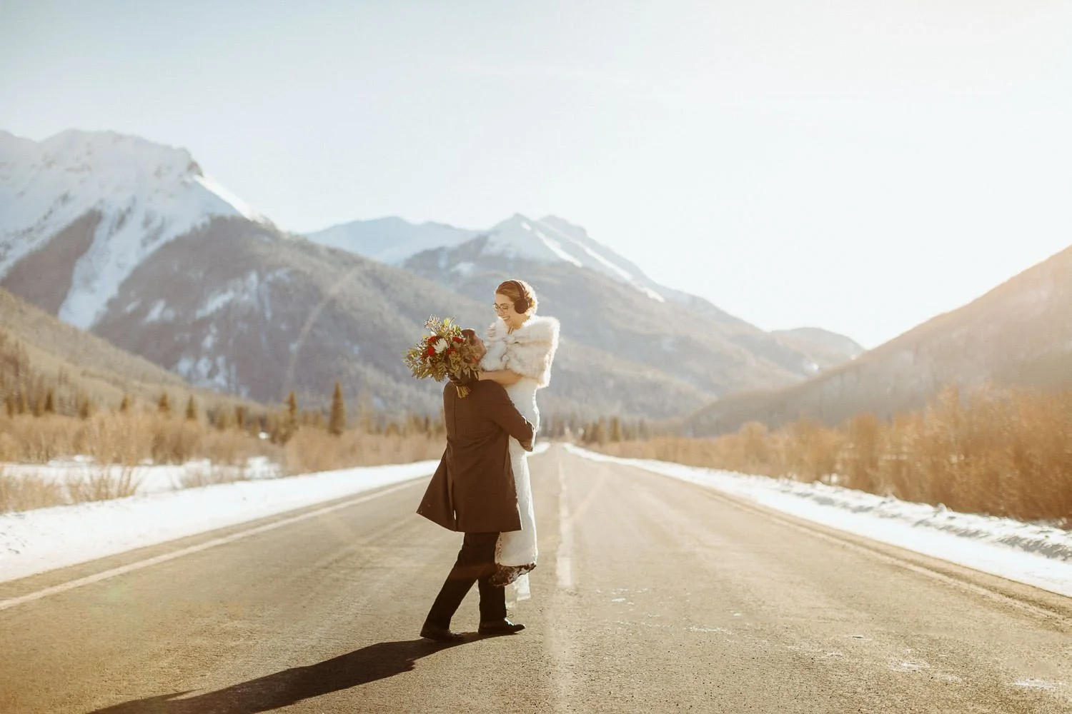 A couple in long coats hugs in the middle of an empty mountain road with snowy peaks behind them