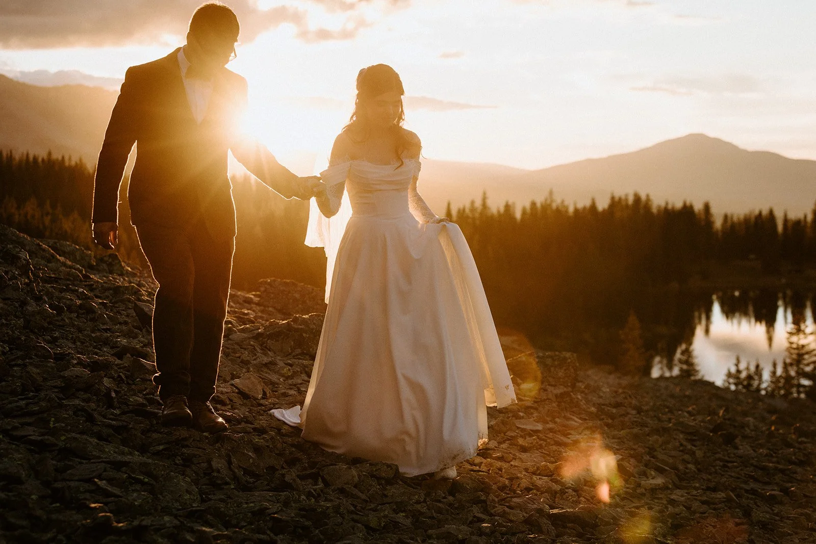 A bride and groom holding hands at Alta Lakes Observatory in Telluride while the sun sets behind them and they walk on rocky crags