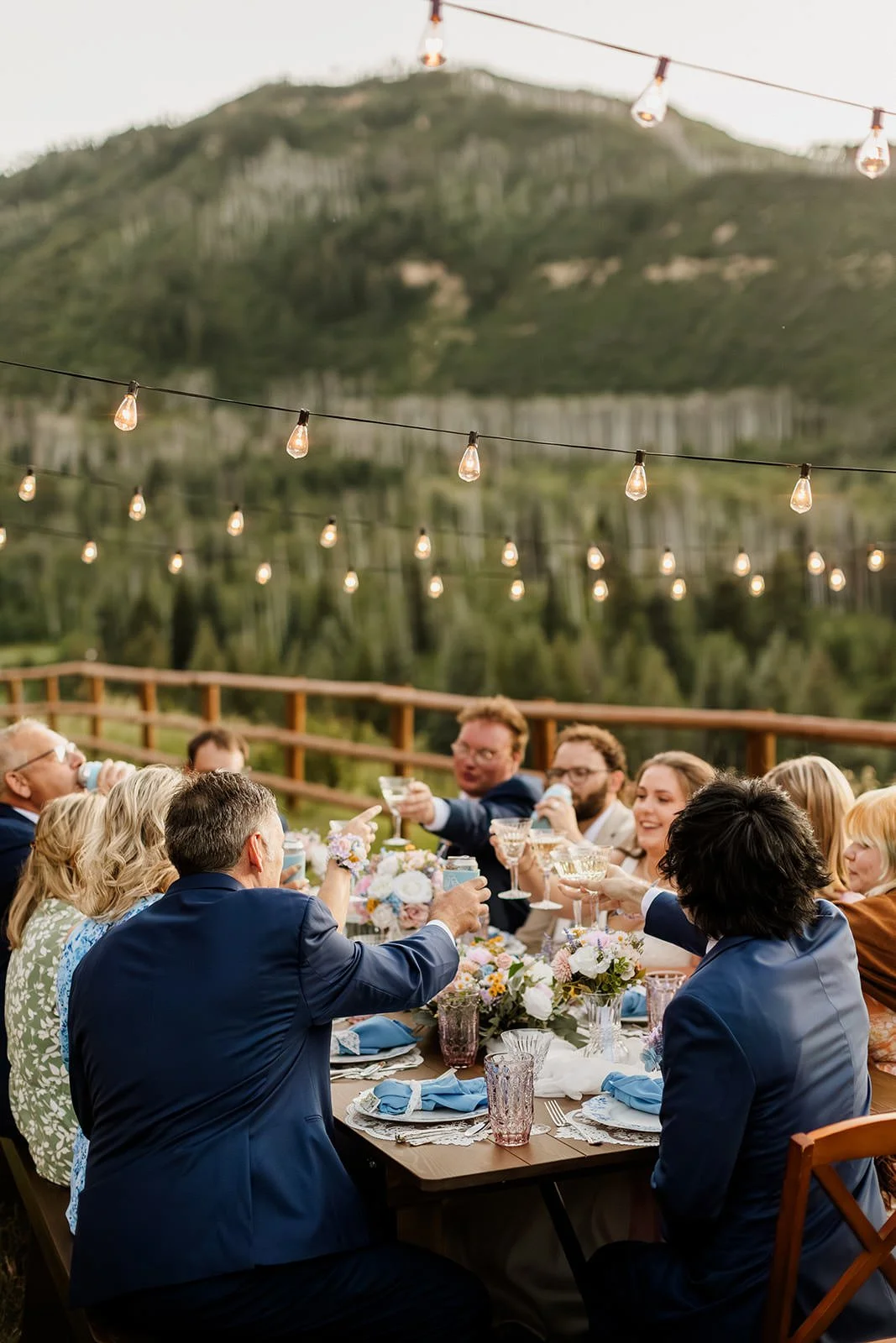 A group of people celebrating at a long outdoor table with string lights overhead, surrounded by greenery and mountains in the background, during daytime.