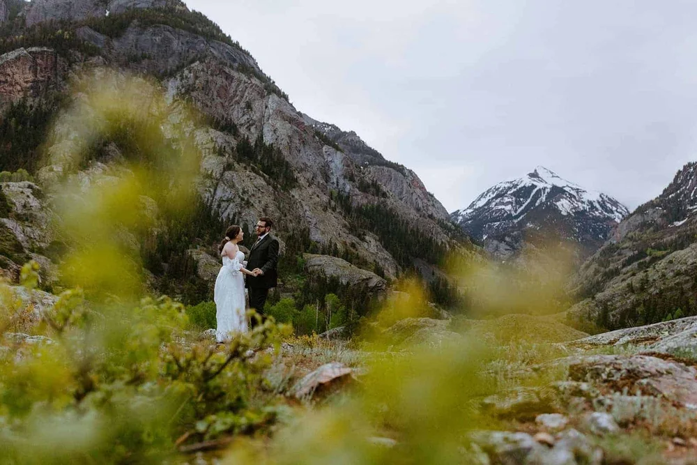 A couple framed by green leaves with snowy San Juan peaks behind them around Ouray, Colorado