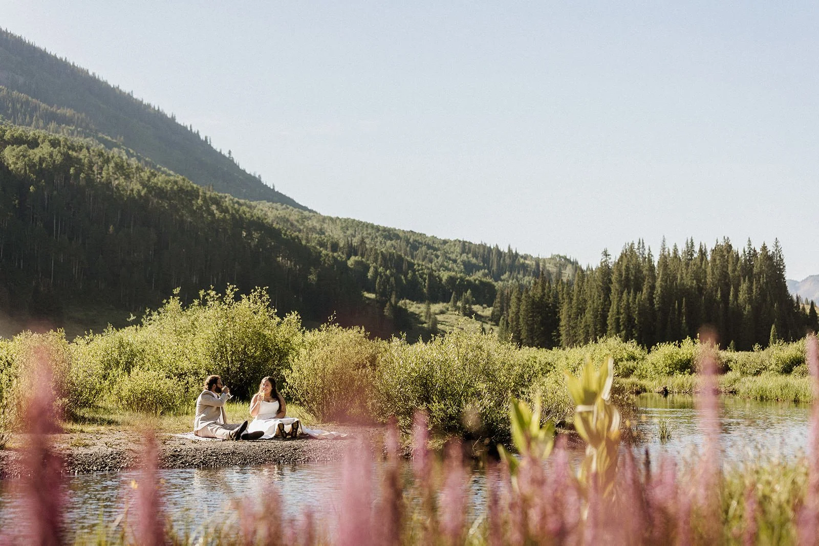 A man and woman sitting on a blanket by a river, having a picnic with a backdrop of green forested mountains under a clear sky.