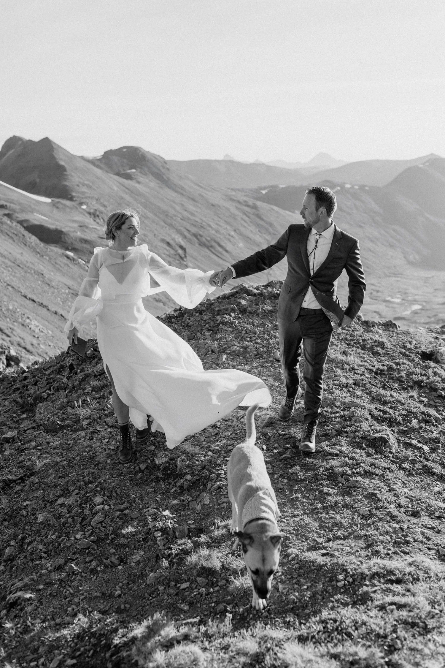 Kimberly Crist in her wedding dress with sheer sleeves, holding hands with her partner on a mountainside