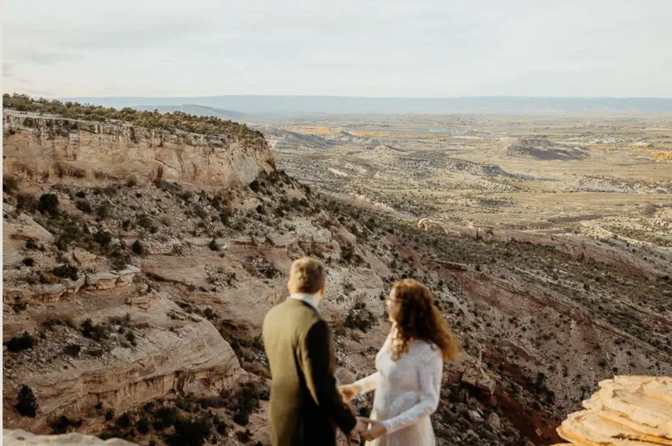 A couple holds hands on a canyon rim near Grand Junction, looking out over red rock and desert