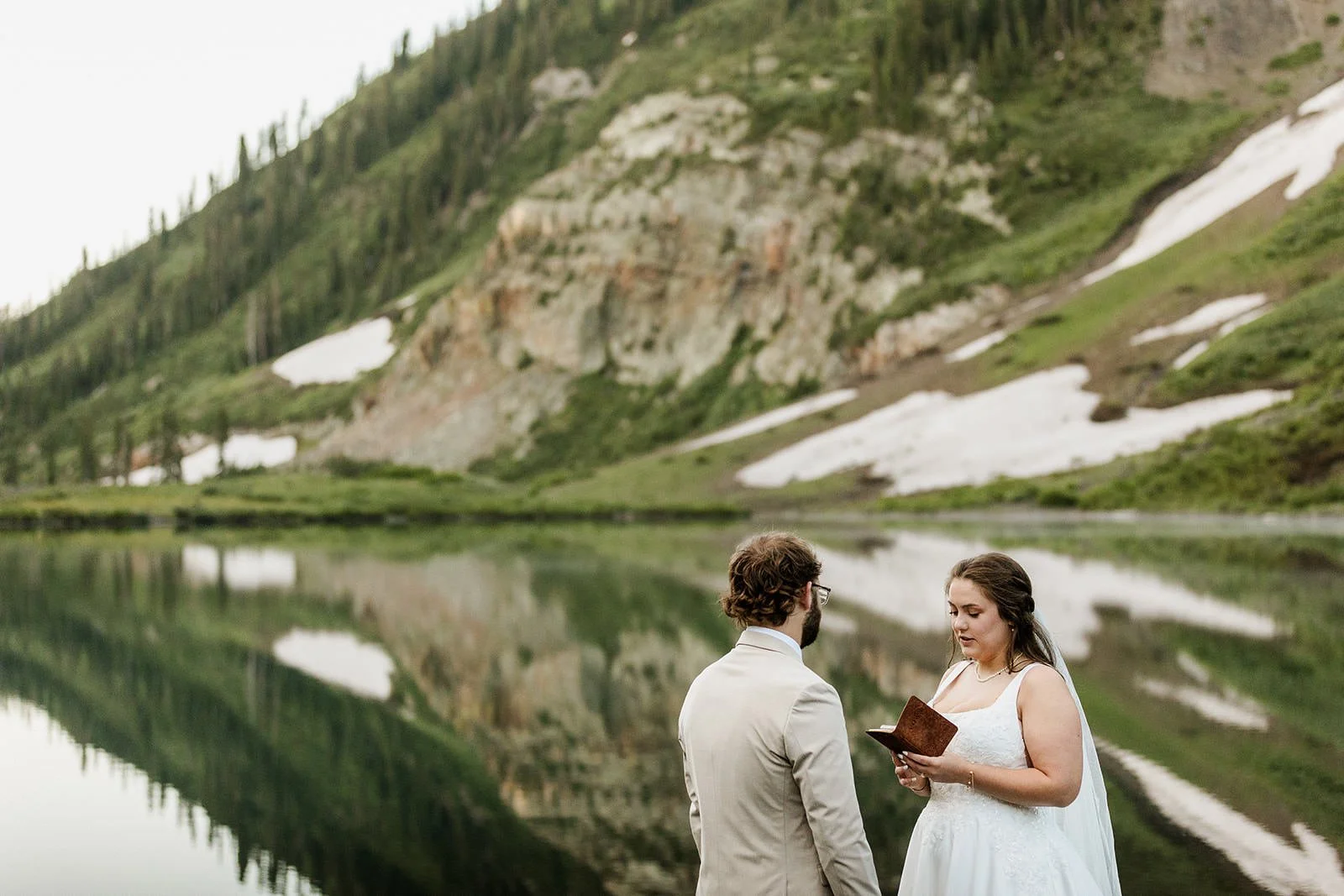A bride and groom standing by a lake with a mountain and trees in the background, the bride holding a small notebook or book.