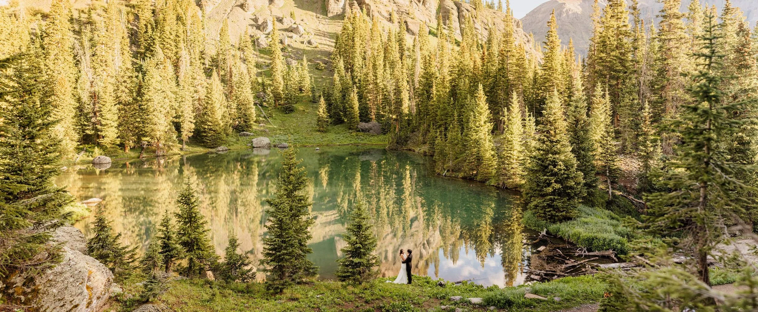 A bride and groom stand on the lake shore of a turquoise alpine lake in Ouray