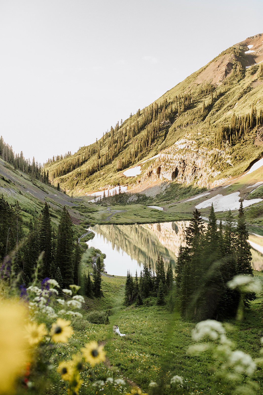 Scenic mountain landscape with a lake, surrounded by evergreen trees and wildflowers, with snow patches on the mountains and a couple sitting on the grassy area by the lake.
