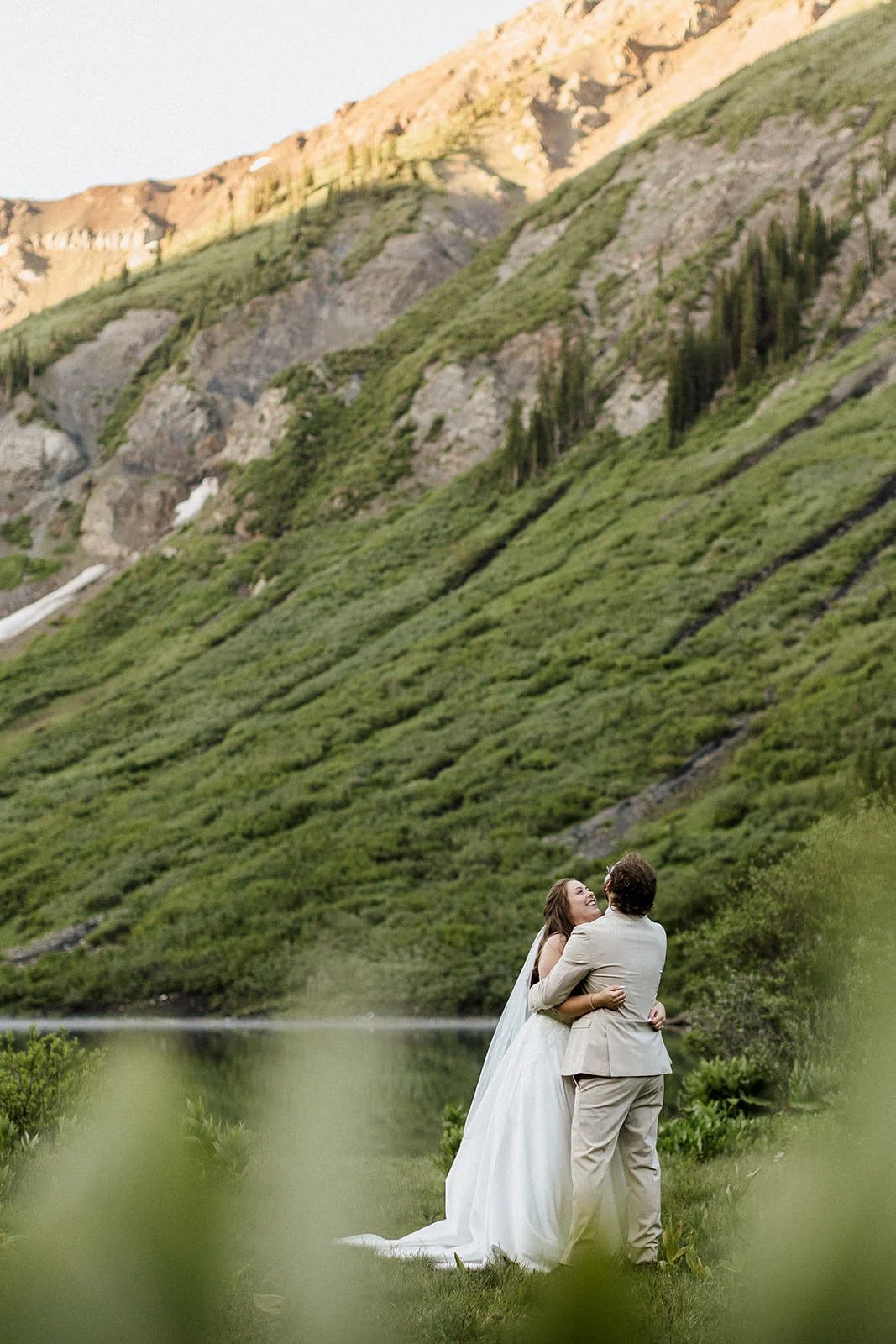A bride and groom embrace near a body of water with a green hillside and rocky mountains in the background, during what appears to be late afternoon or early evening.