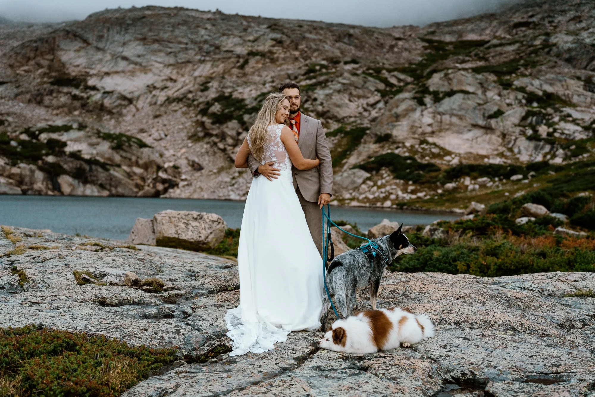 Bride resting her head on her groom's shoulder by an alpine lake, their two dogs on leashes at their feet