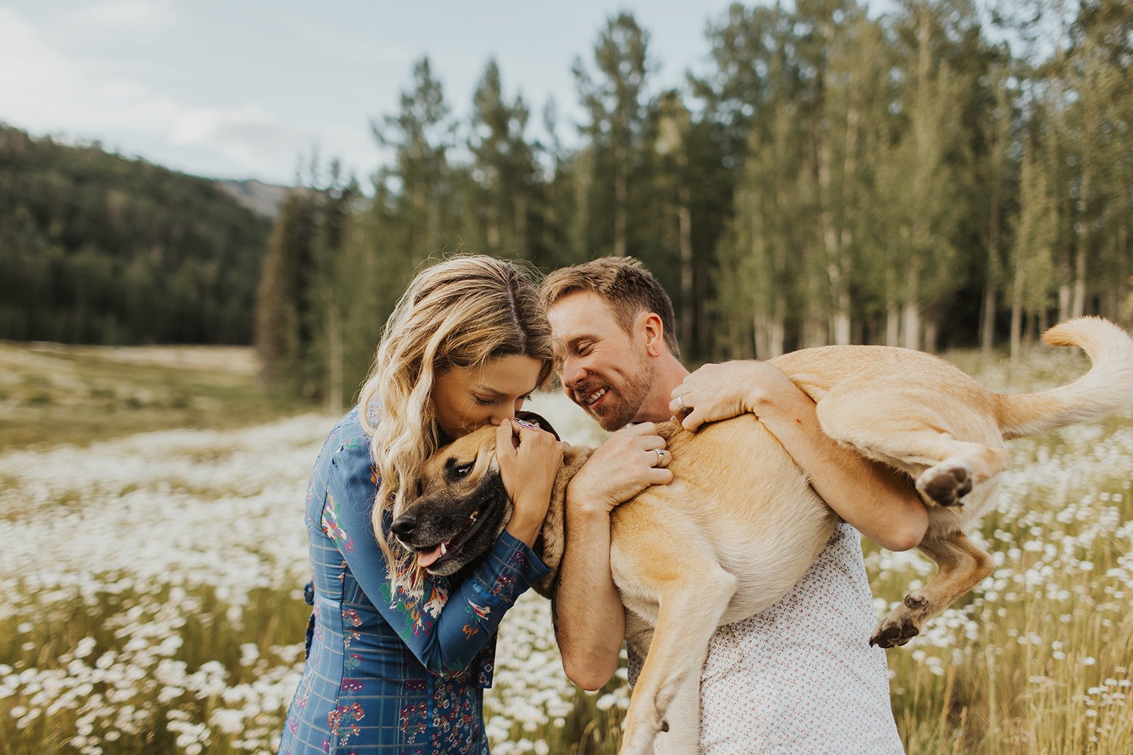 A couple hugging and playing with a dog in a field of white flowers, with a background of trees and mountains.