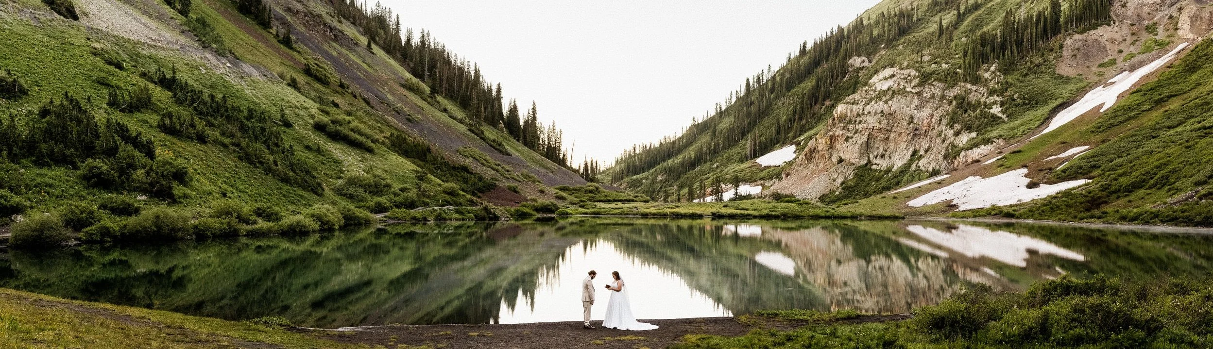 A scenic mountain lake with lush green slopes and snowy patches, reflecting the sky and surrounding mountains. Two women in wedding dresses are standing near the shore, facing each other, holding hands in a picturesque outdoor setting.