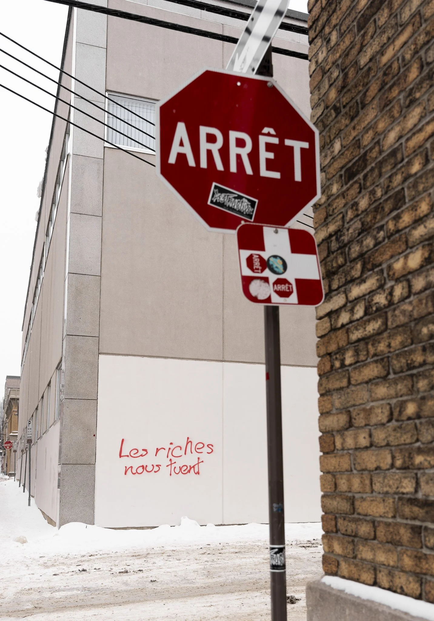 Un panneau d'arrêt rouge avec le mot 'ARRÊT' en français, fixé sur un poteau dans une zone urbaine en hiver, avec un mur en brique à droite et un bâtiment moderne en arrière-plan. Sur le mur blanc derrière, une inscription en rouge dit 'Les riches no