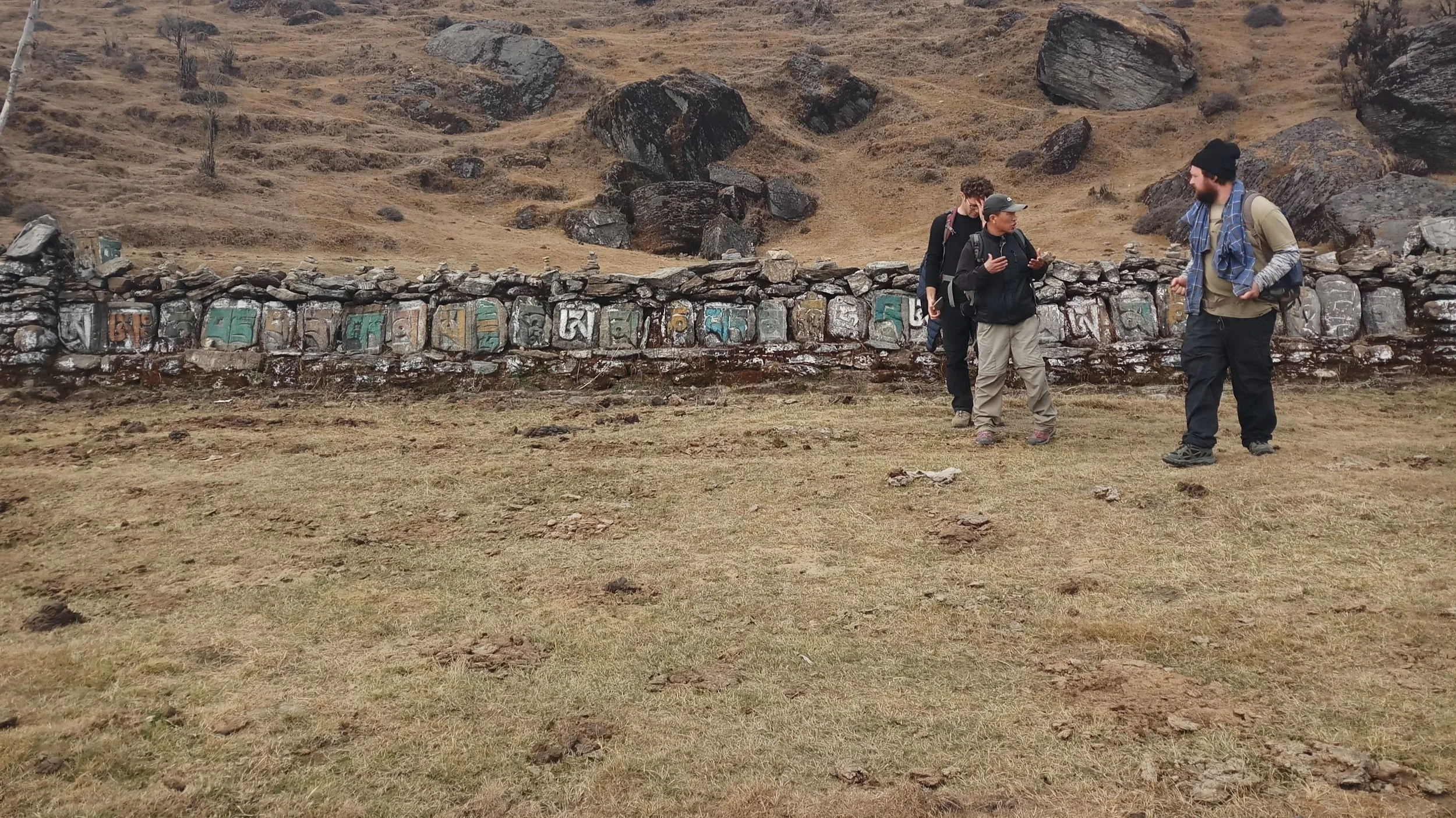 Trois personnes en randonnée discutant devant un mur en pierre avec des peintures colorées, dans un paysage de montagne avec des rochers et de l'herbe sèche.