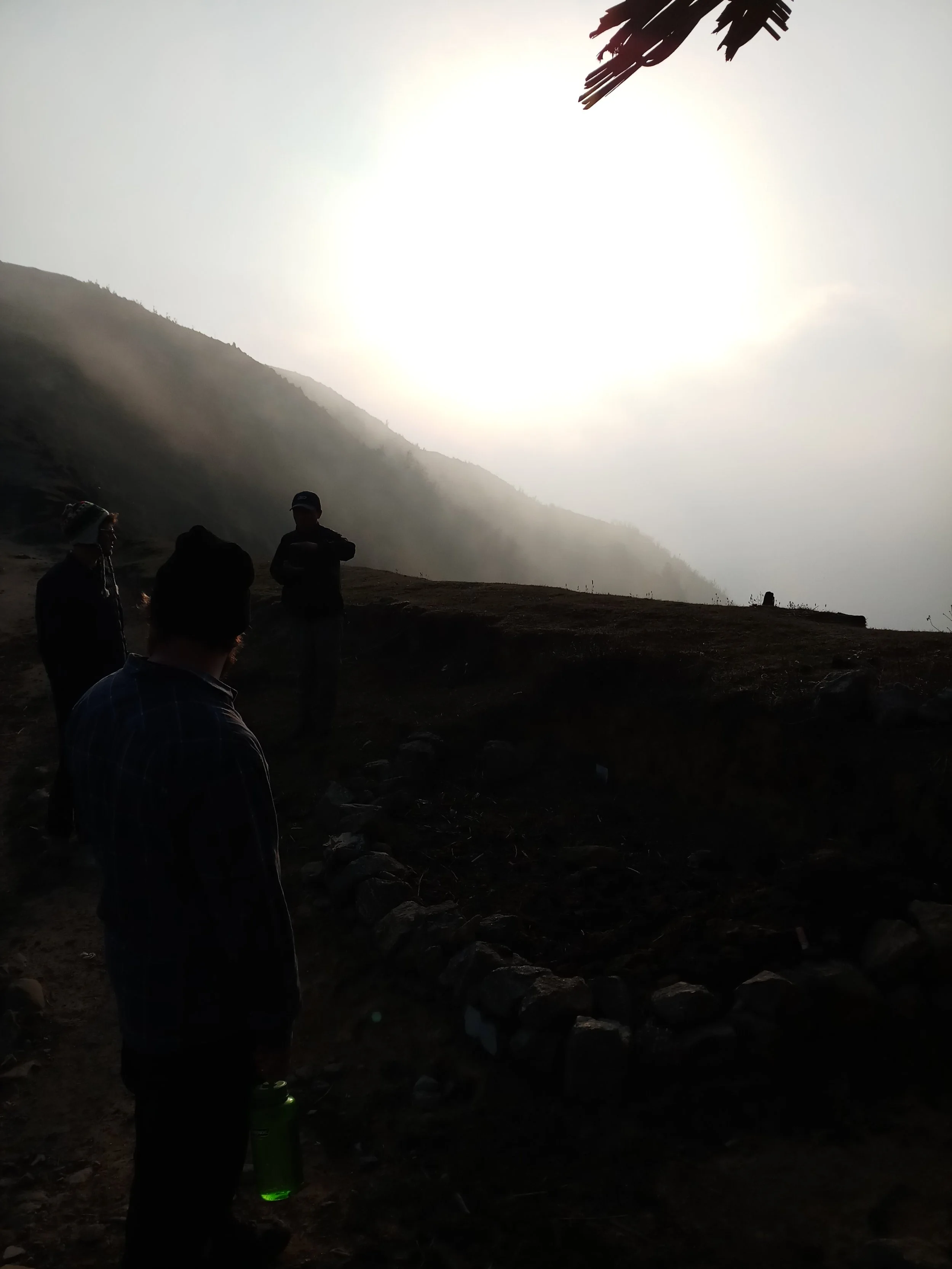 Trois personnes regardent une opération de fouilles archéologiques dans un paysage montagneux, avec le soleil brillant en arrière-plan.