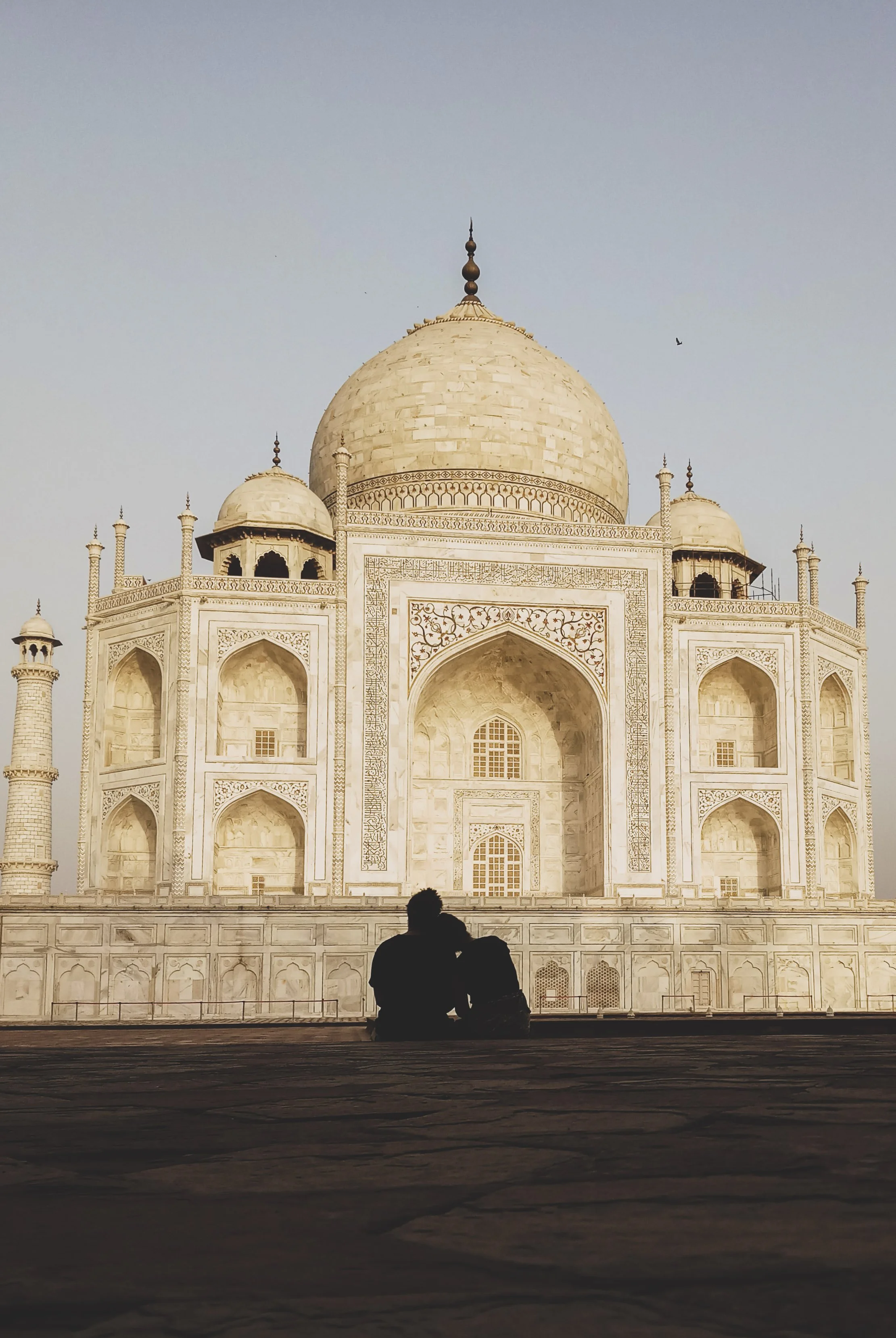 Silhouettes de deux personnes assises devant le Taj Mahal, monument en marbre blanc en Inde, avec un ciel clair en arrière-plan.