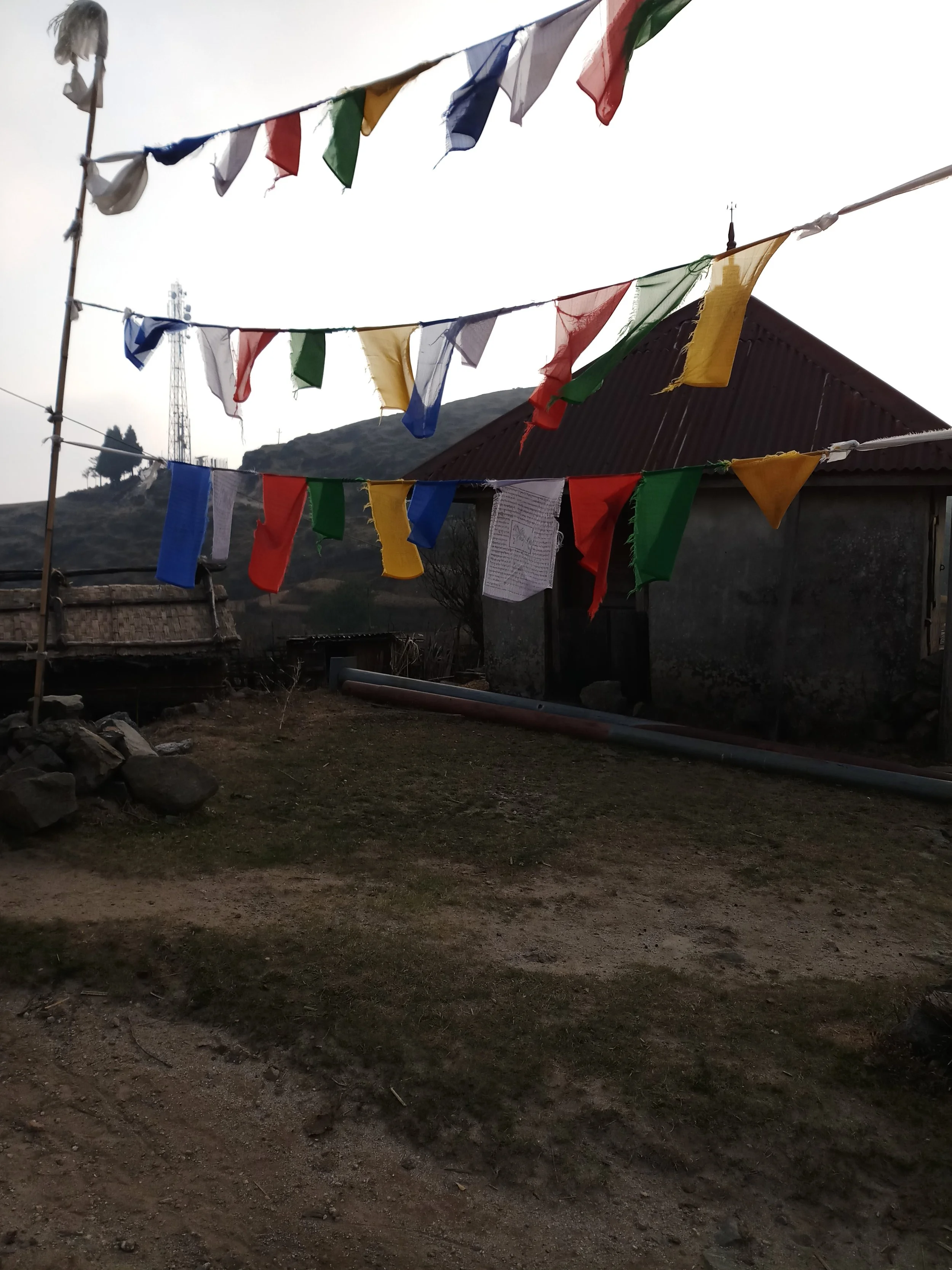 Drapeaux de prière colorés suspendus en extérieur, avec une maison en arrière-plan, sur un terrain en pente avec quelques pierres.