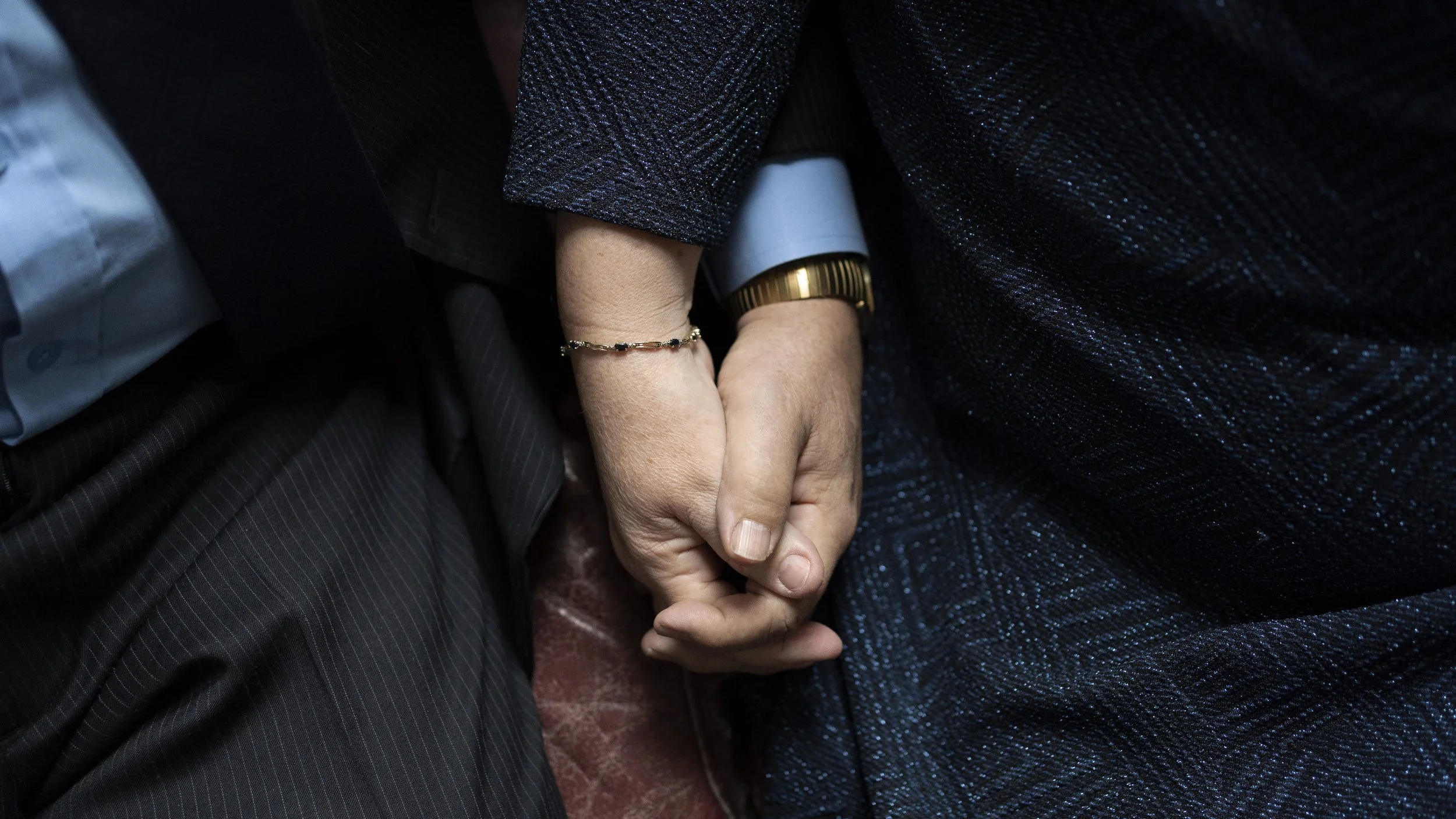 Gros plan sur deux mains jointes, une main d'homme et une main d'une femme, avec la femme portant une montre en or et un bracelet délicat, les deux personnes portent des vêtements sombres