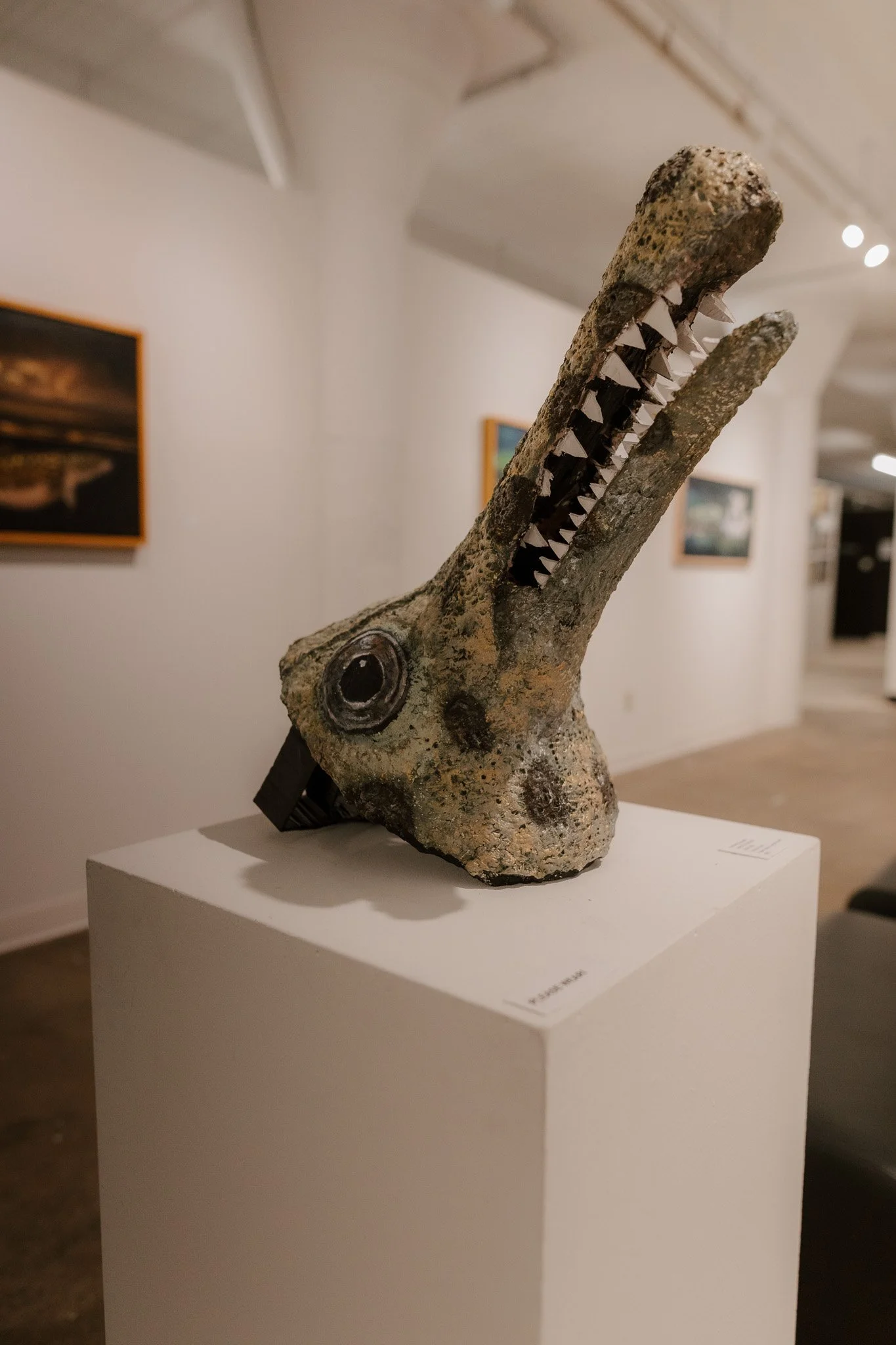 Art sculpture resembling a crocodile head with a long snout, sharp teeth, and textured surface, displayed on a white pedestal in an art gallery.