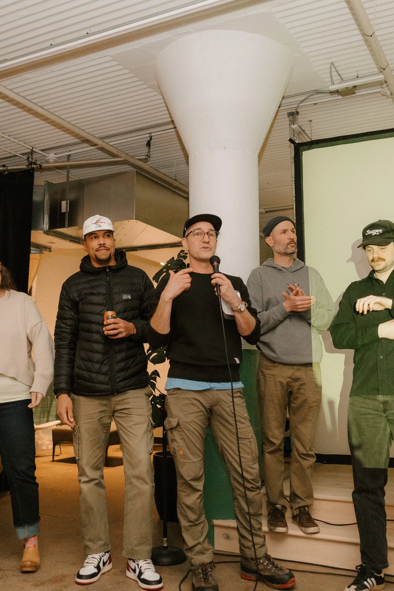 Group of four men standing indoors, with one man in the center holding a microphone and speaking, while the others listen. They are casually dressed, and the setting appears to be a presentation or event space with a large screen behind them.