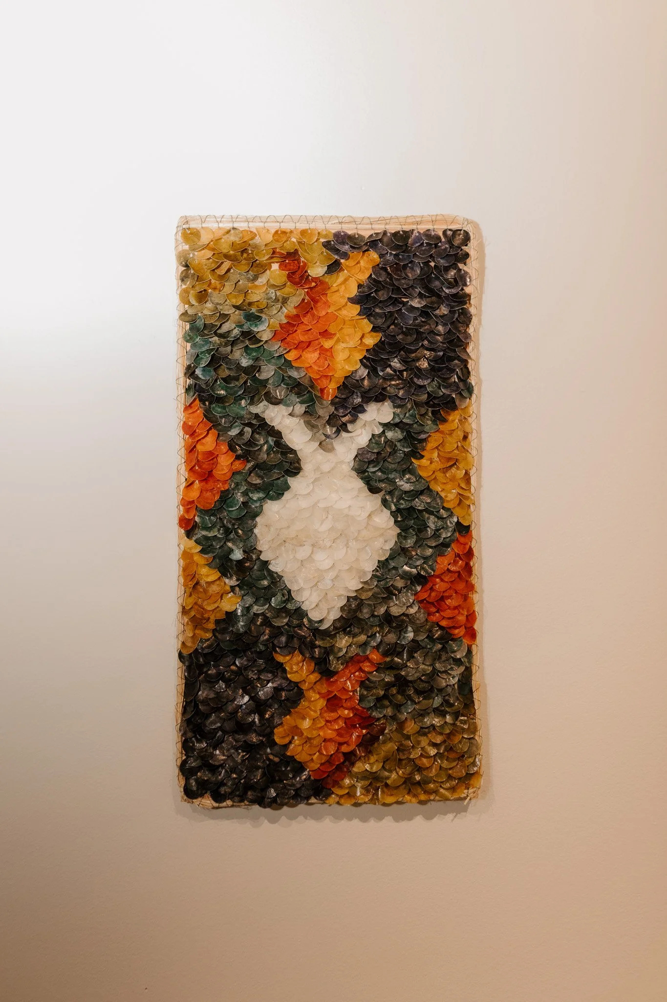 A colorful mosaic artwork on a rectangular canvas with a black border, composed of numerous small shells arranged to form an abstract pattern with shades of orange, black, white, and green.