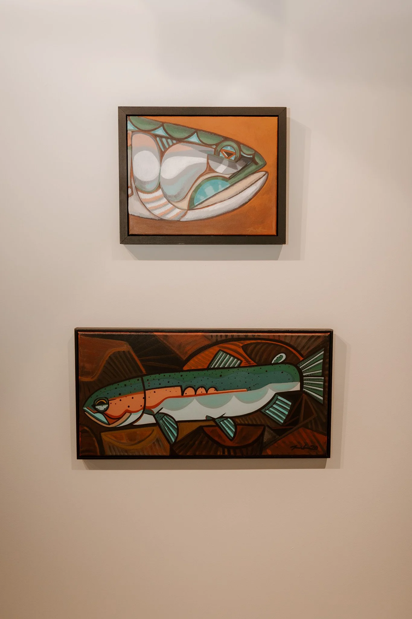 Two framed abstract paintings of fish hanging on a white wall, one above the other.