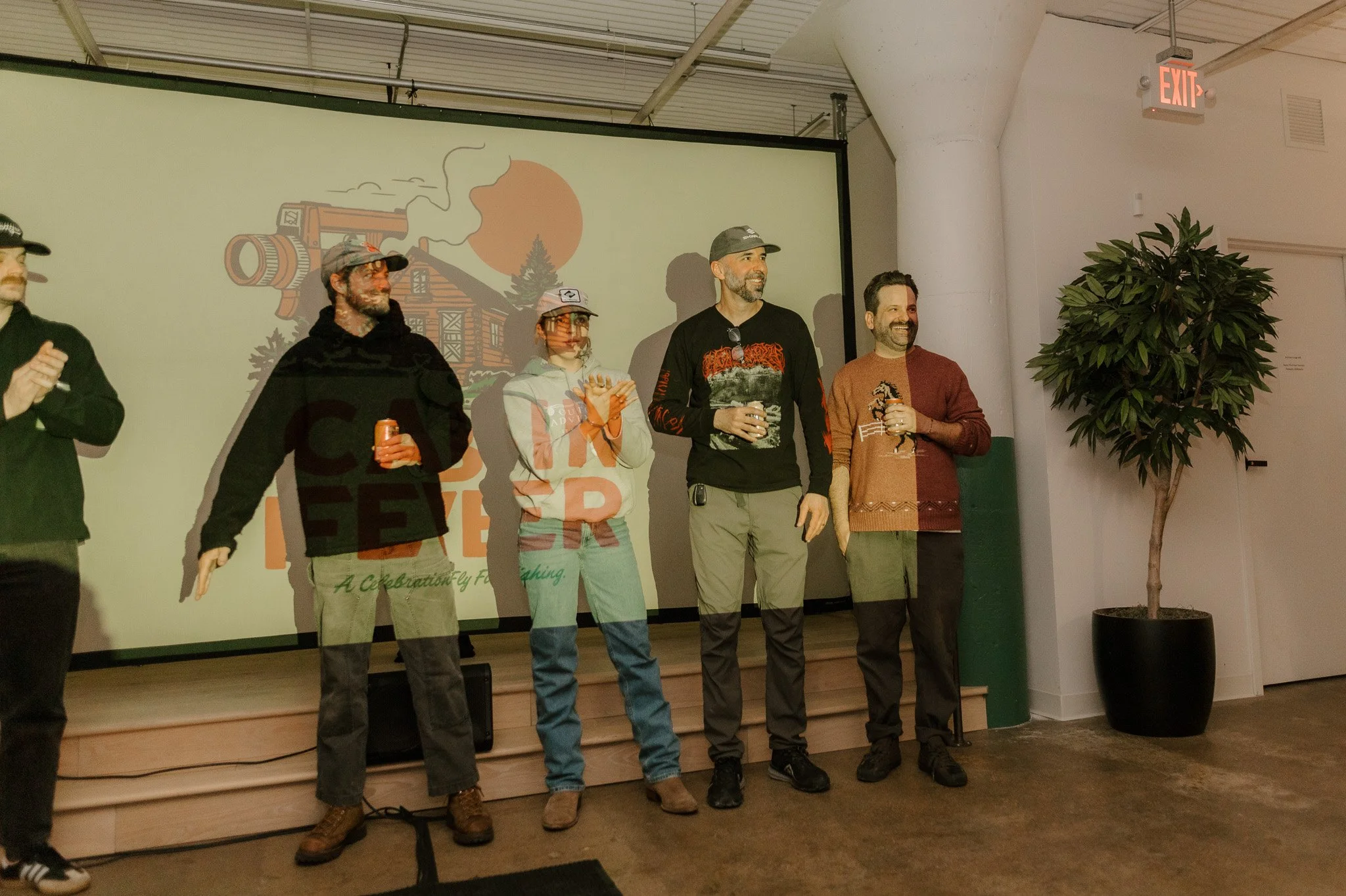 Group of five people standing on a stage holding drinks, in front of a screen with a logo and outdoor scene, with a potted plant and exit sign in the background.