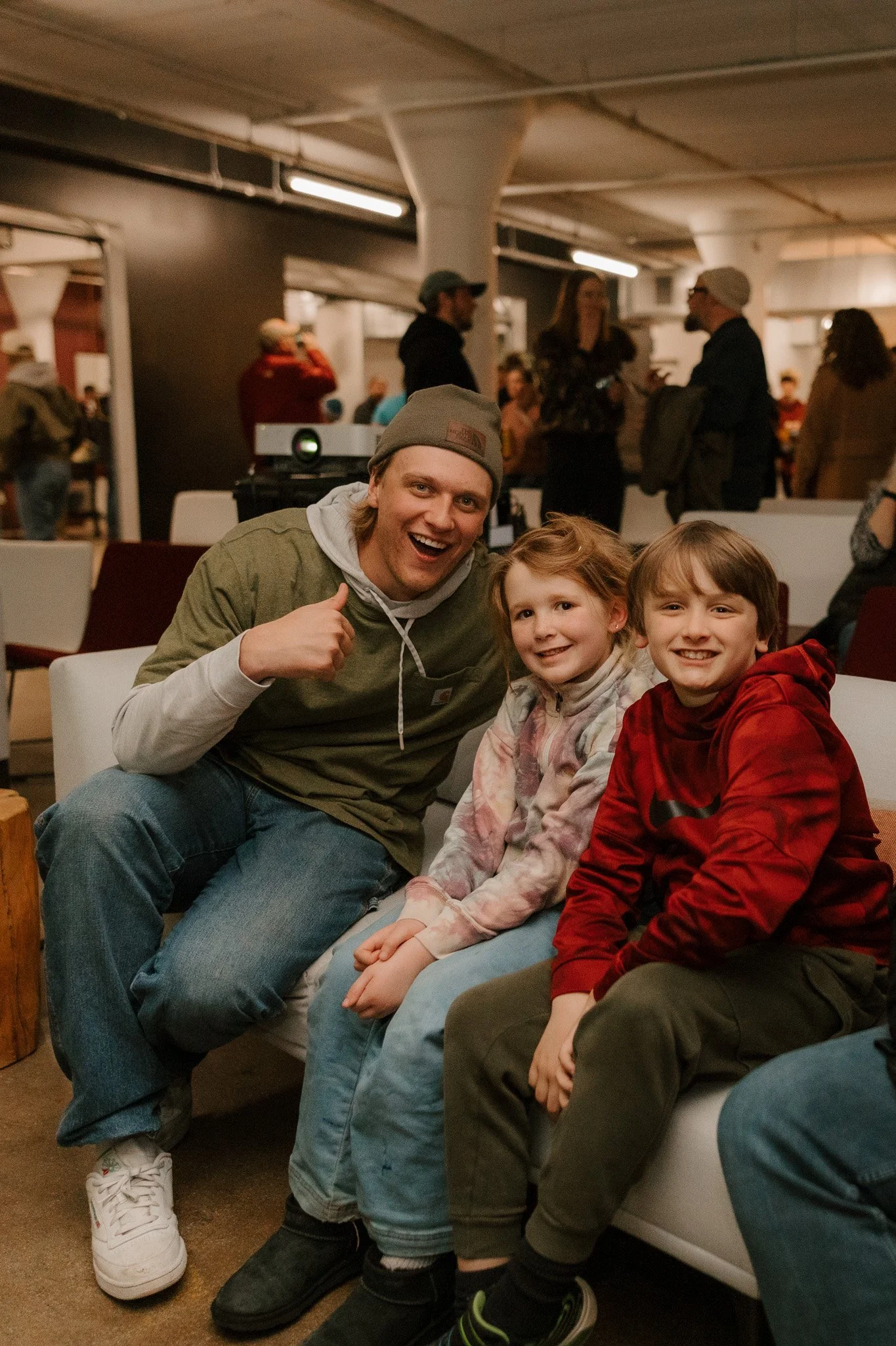 A group of three people, one adult man and two children, sitting on a couch at a social event or gathering. The man is giving a thumbs-up and smiling, wearing a gray beanie and casual clothing. The children are sitting beside him, smiling at the came
