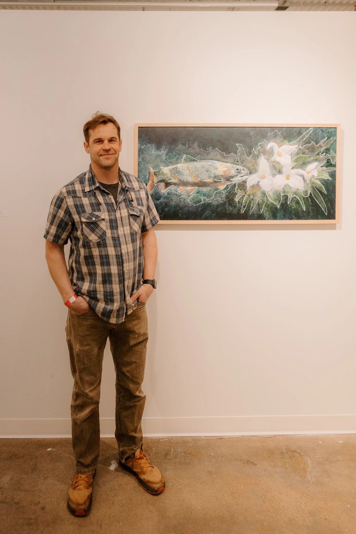 A man with short brown hair, wearing a plaid shirt, khaki pants, and hiking boots, stands with his hands in his pockets in front of a painting of a fish and white flowers at an art gallery.