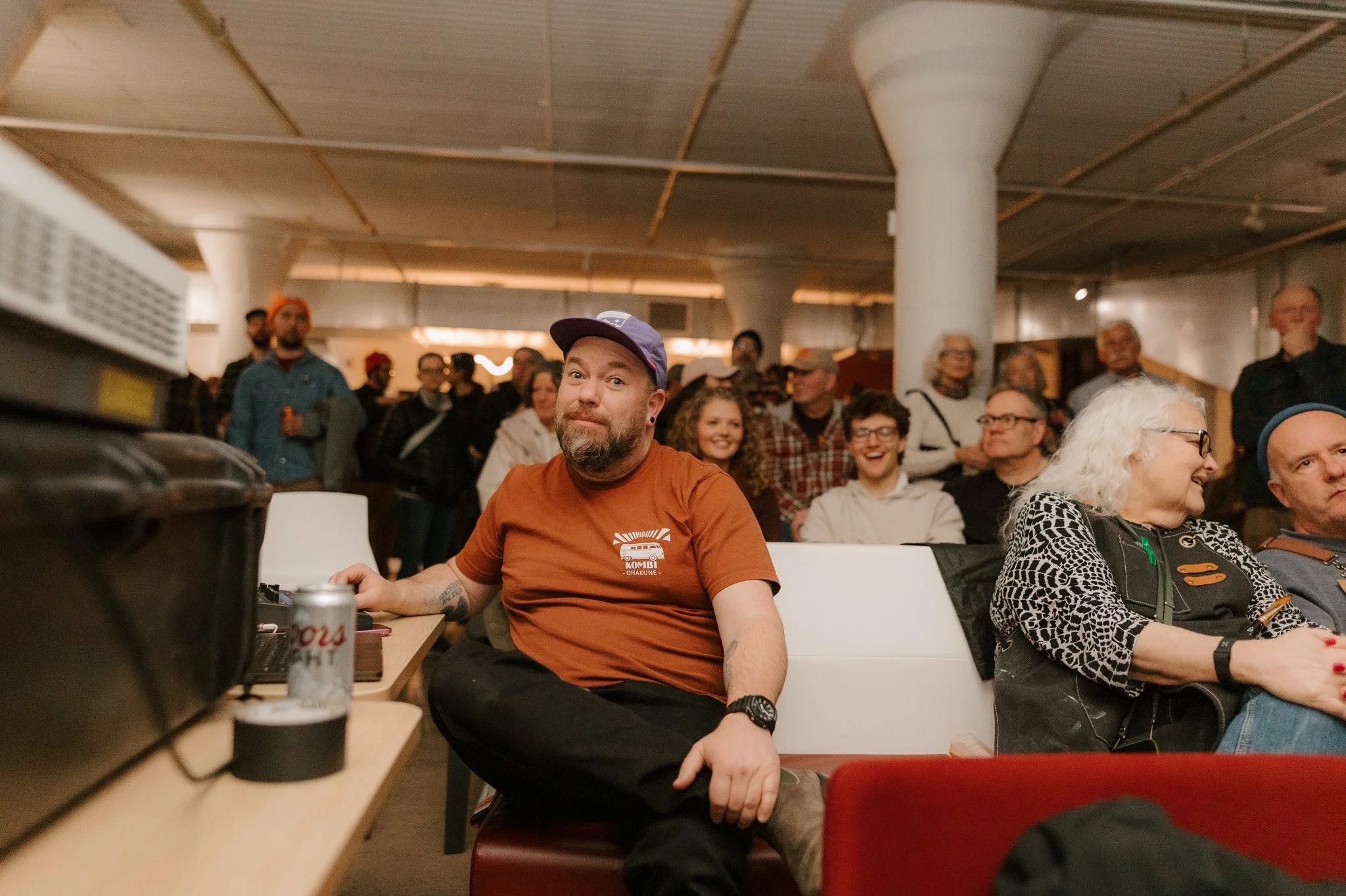 Audience watching a presentation or event in an indoor venue, with a man in an orange shirt and purple cap seated in the front row, holding a can of Coors Light, and a group of people laughing and smiling in the background.