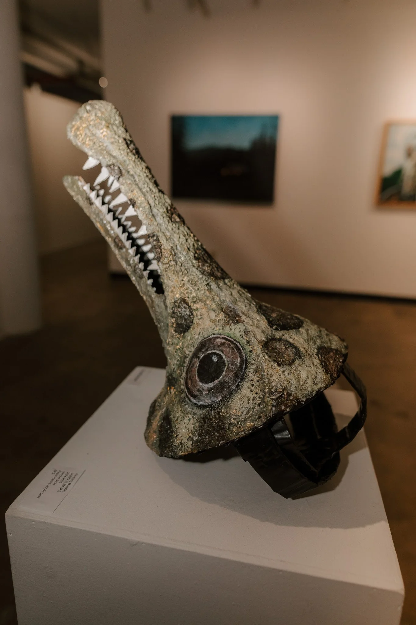 Sculpture of a creature with a crocodile or dinosaur head and an open mouth, showing sharp teeth, displayed on a white pedestal in an art gallery.