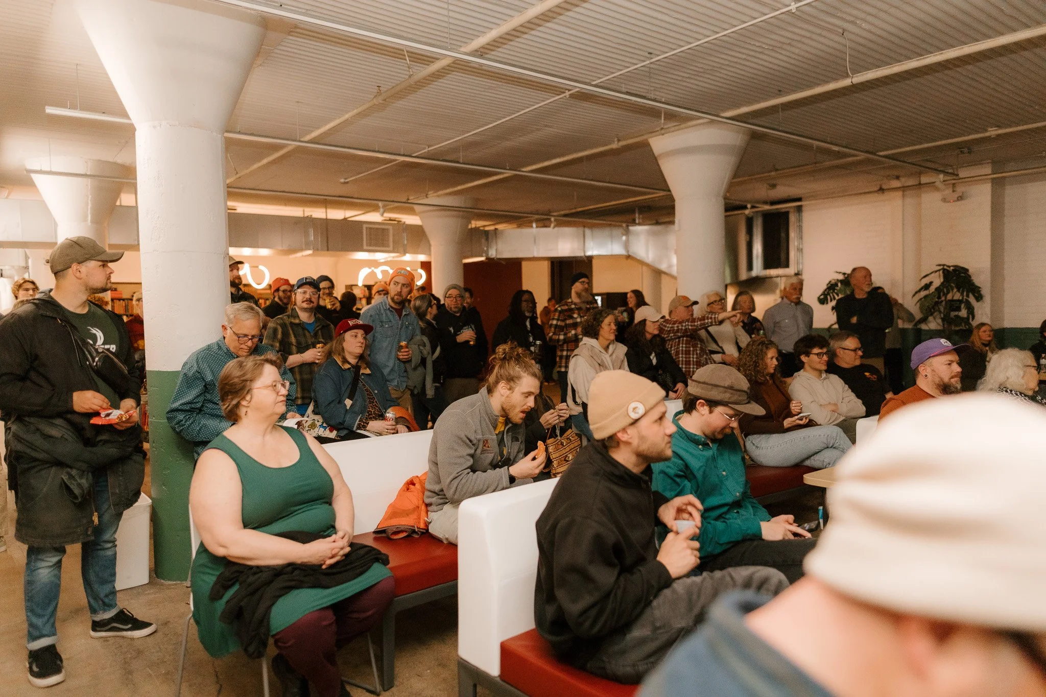 An indoor event with a large group of people seated and standing, listening to a presentation or speaker. The setting has white brick walls, exposed ceiling pipes, and large columns.