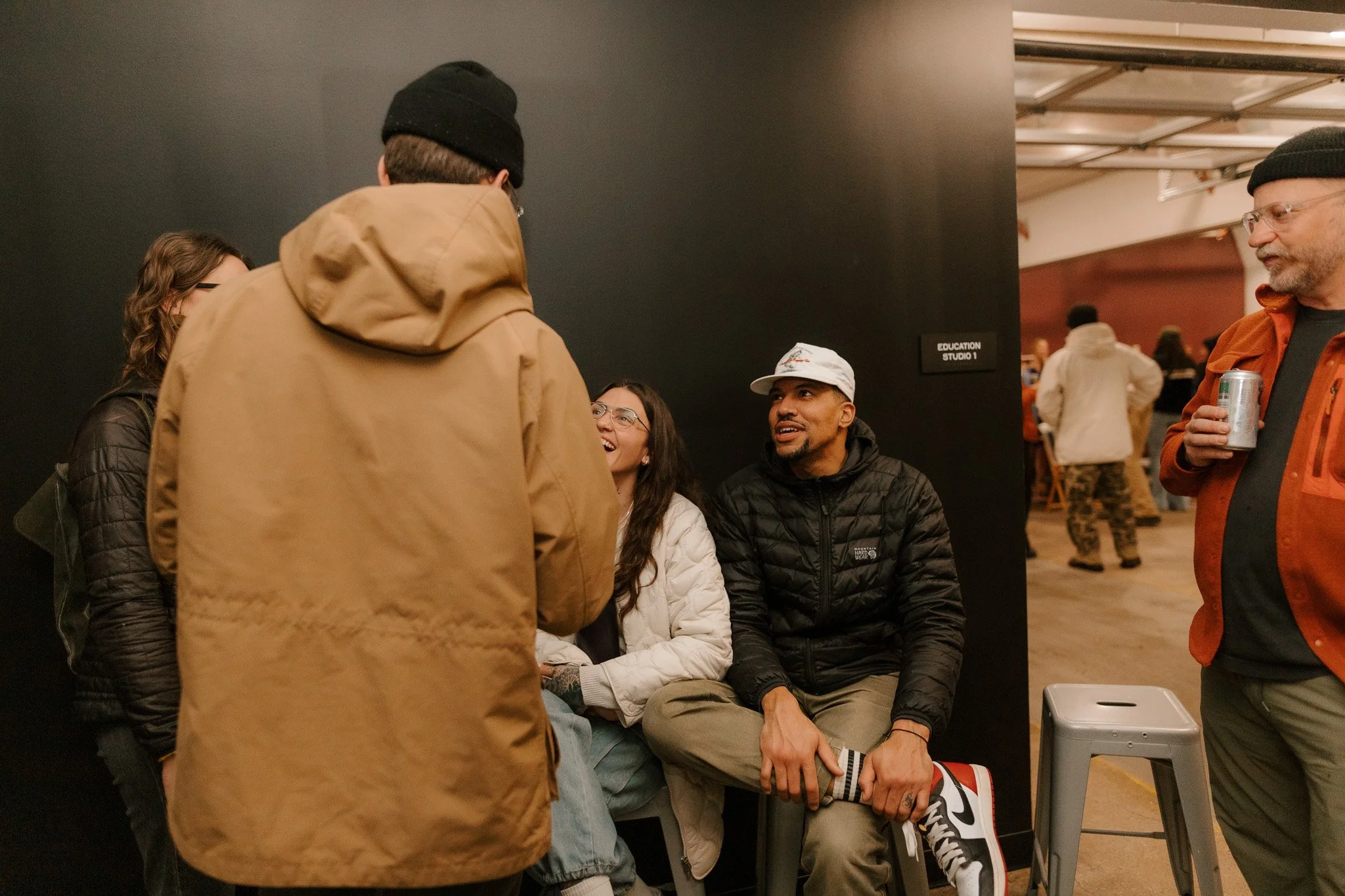 A group of five people conversing in an indoor setting, with four seated and one standing. The seated individuals include a woman with glasses and long dark hair, and a man with a baseball cap and a black puffer jacket. The standing person is a man i