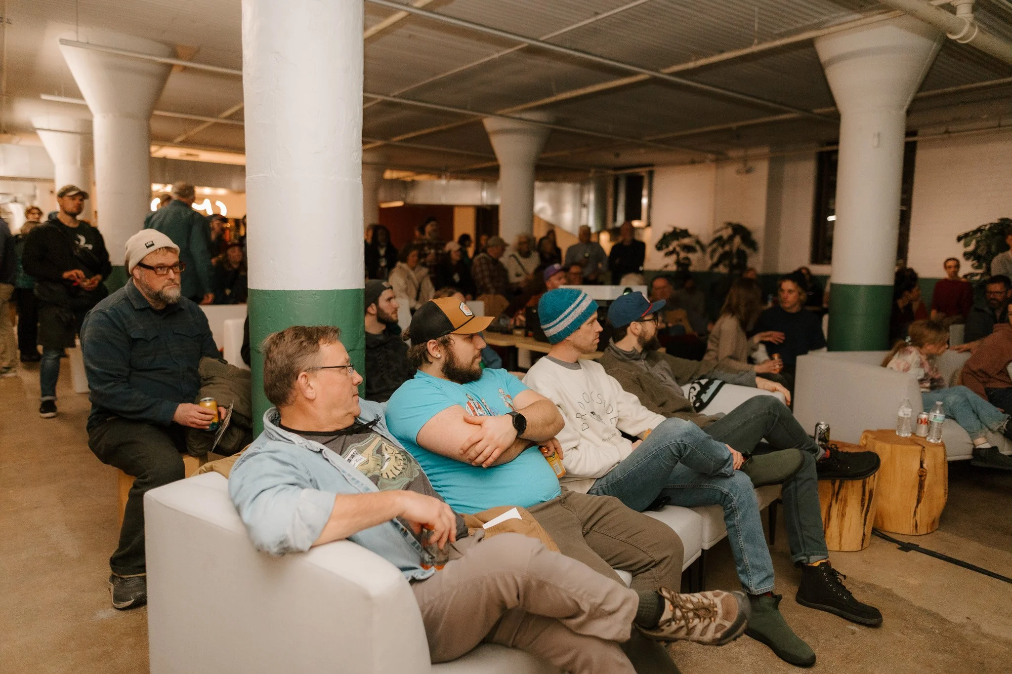 People sitting and standing in a spacious indoor event space with green and white columns, watching an event or presentation.