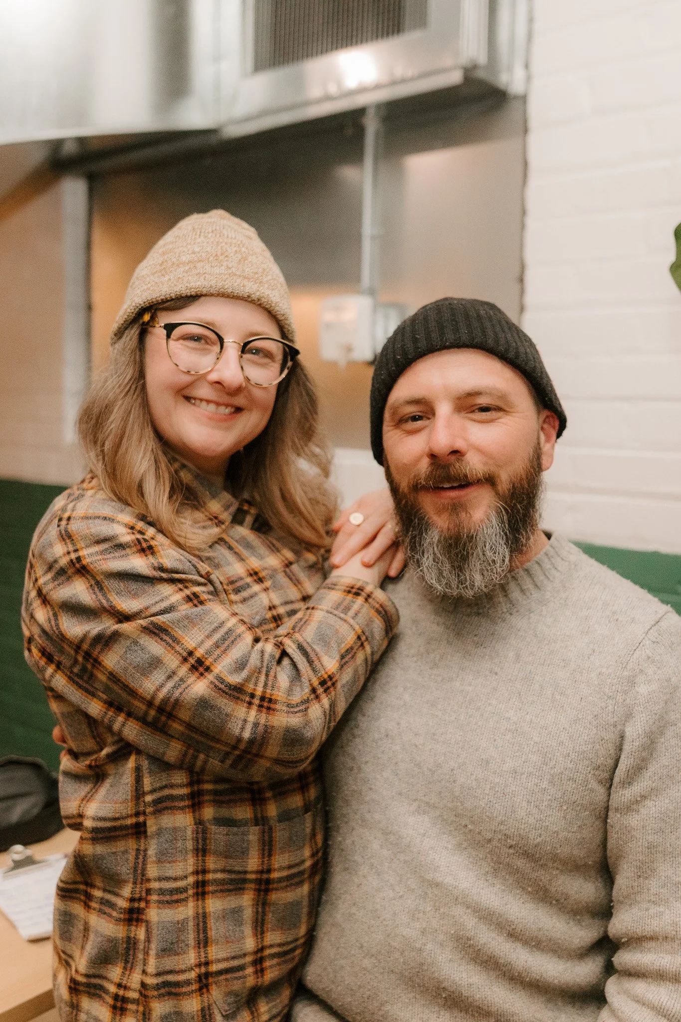 A smiling woman and man wearing knit hats in a cozy indoor setting.