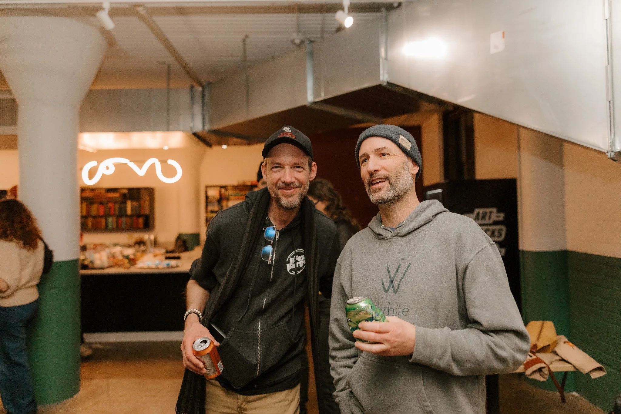 Two men standing indoors, smiling and holding beverages, in a casual setting with bookshelves and a neon sign in the background.