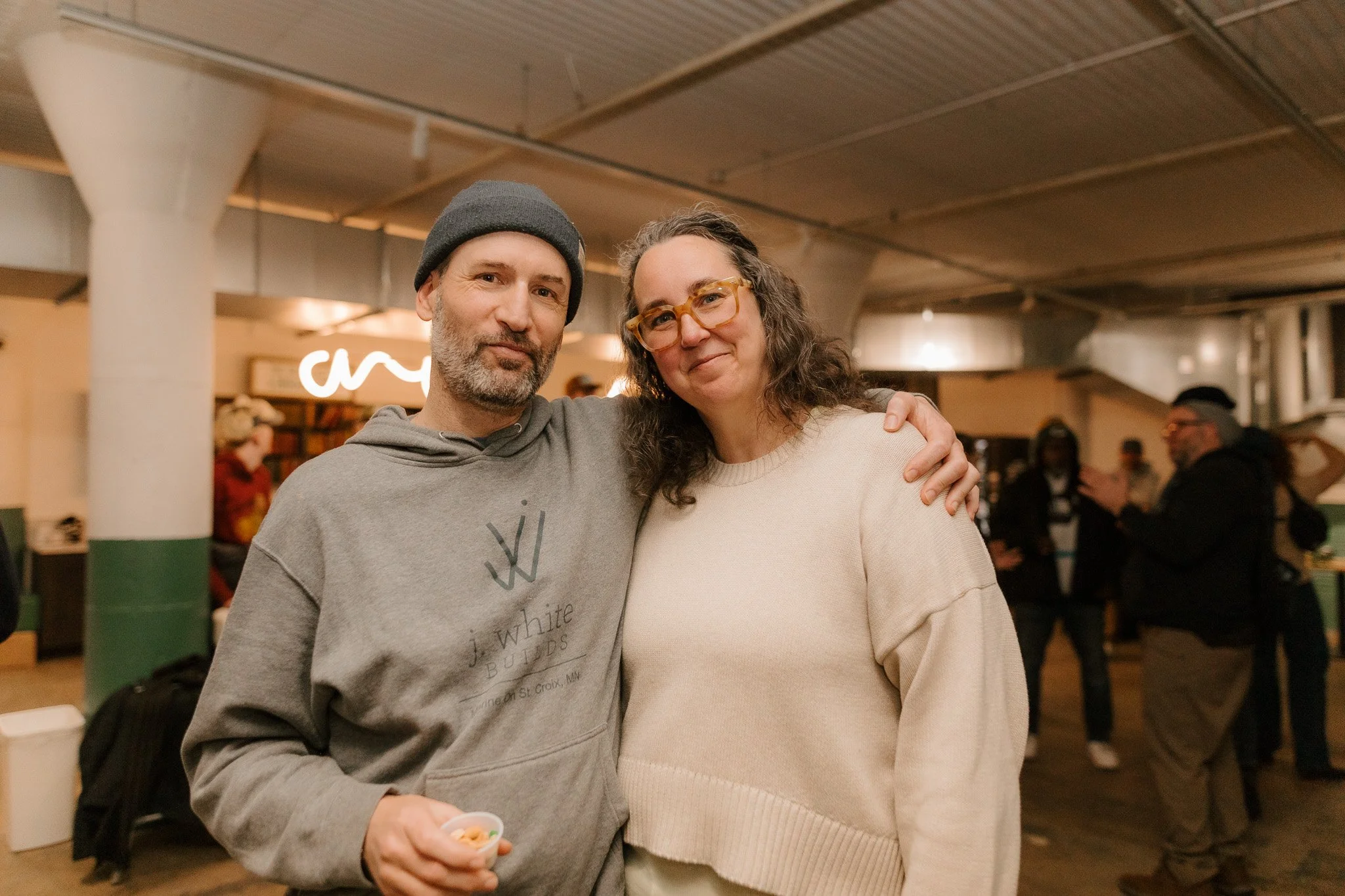 A man and a woman posing together in a social setting, with the man wearing a gray hoodie and beanie, and the woman wearing a cream sweater and glasses.