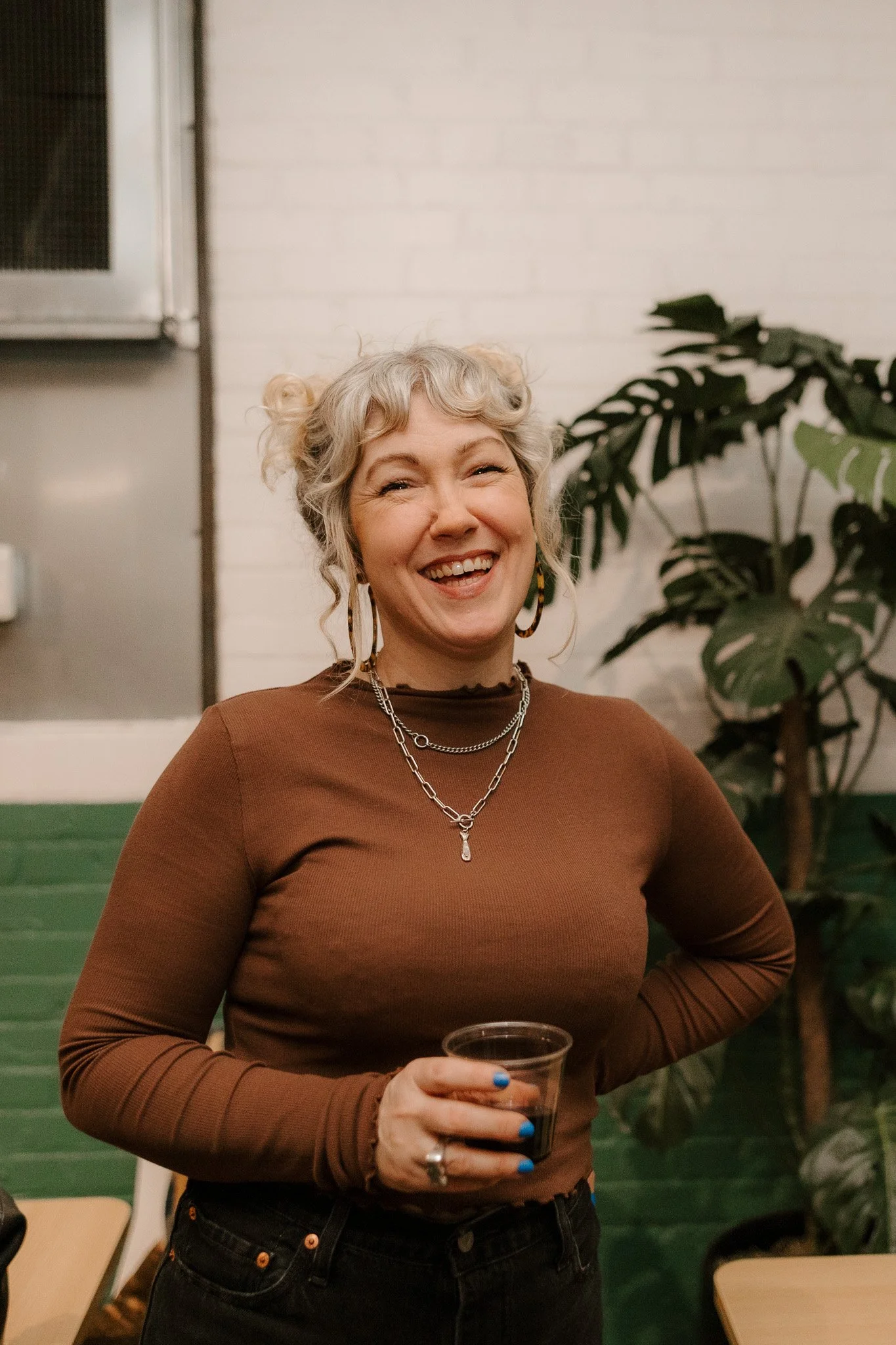 A smiling woman with curly gray hair, wearing a brown long sleeve shirt, multiple necklaces, and colorful jewelry, holding a clear cup with dark liquid, standing in front of a green and white brick wall with a plant on her right.