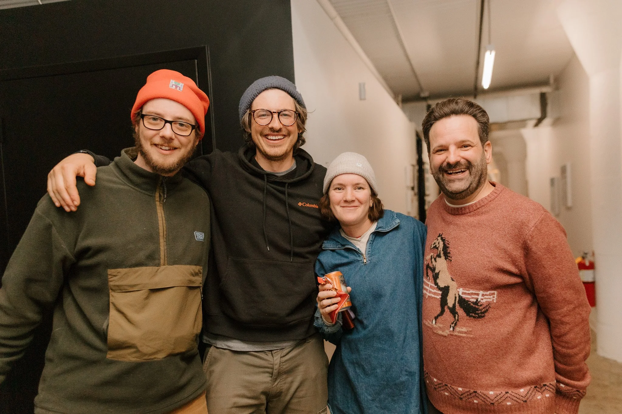 Four friends standing together indoors, smiling, wearing casual winter clothing including hats and sweaters, with arms around each other.