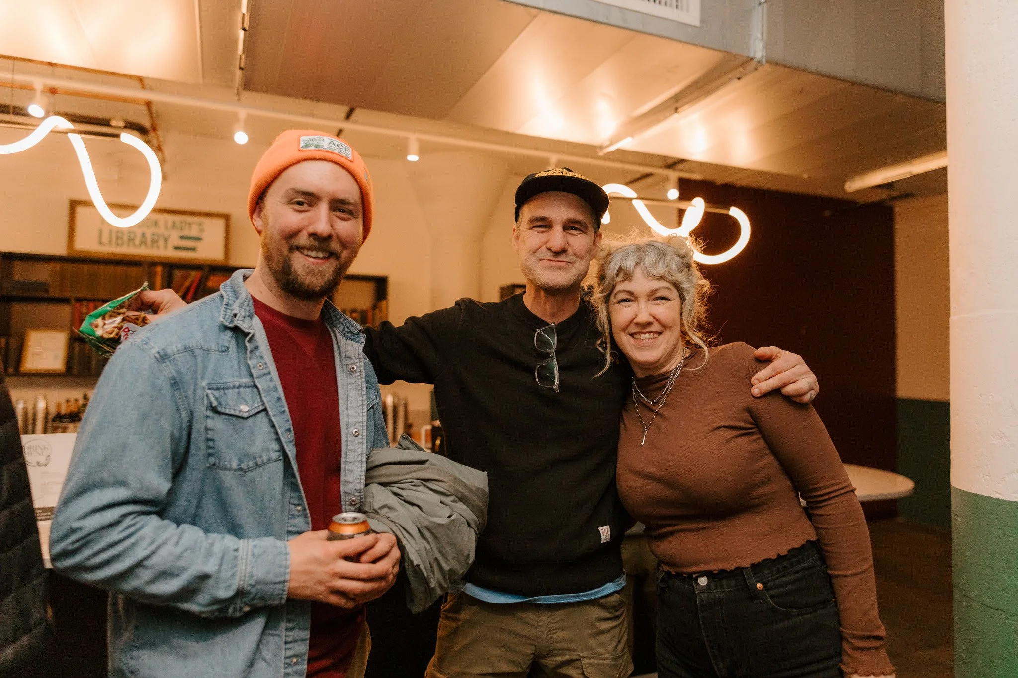 Three people smiling and posing together indoors. The person on the left is a man with a beard, wearing a pink beanie and denim jacket, holding a drink. The person in the middle is a man in a black shirt and cap, with glasses hanging from his neck. T