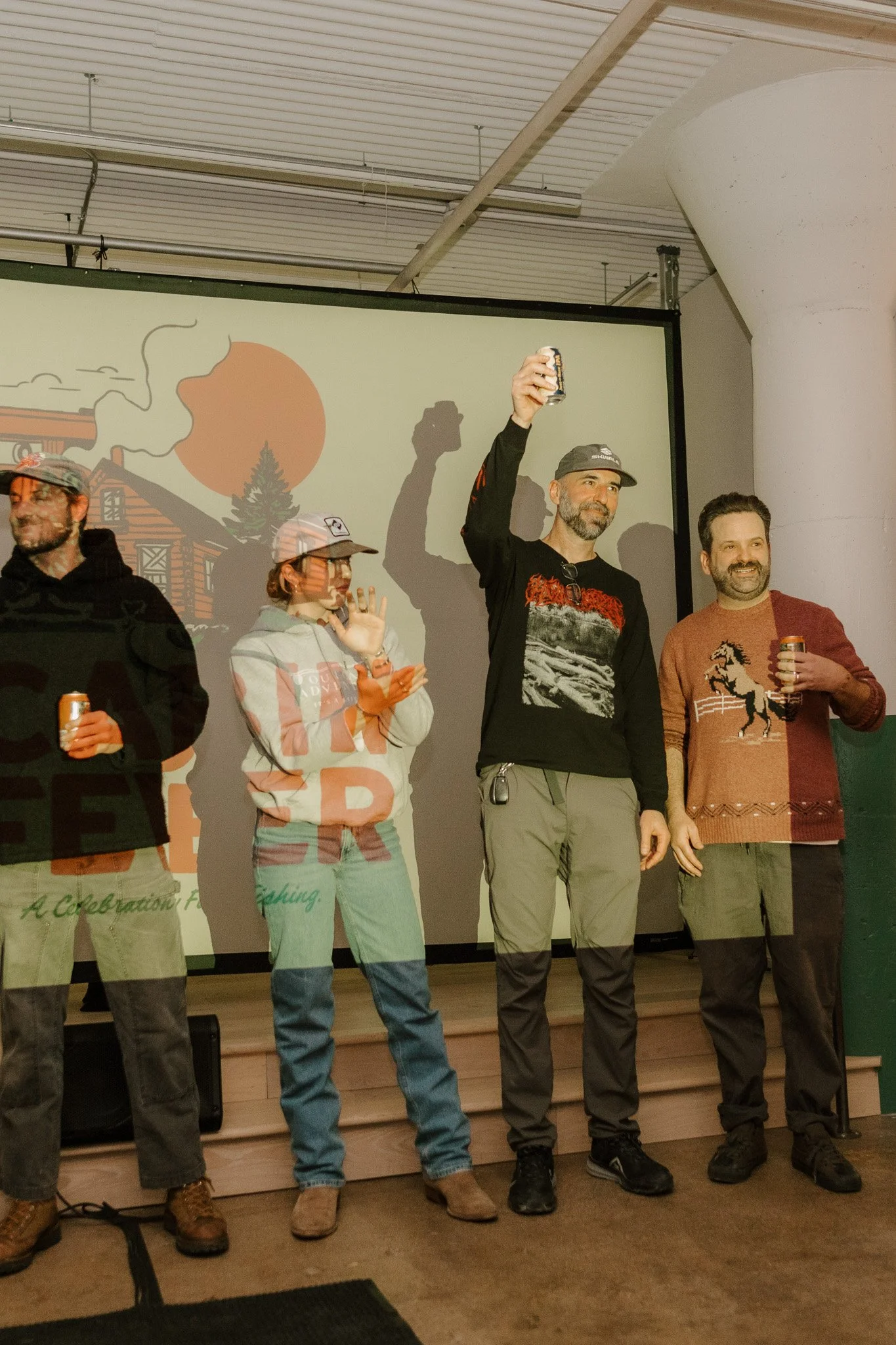 Four people standing in front of a screen, holding drinks, at a celebration or gathering. One person is raising their drink, wearing outdoor or camouflage clothing.