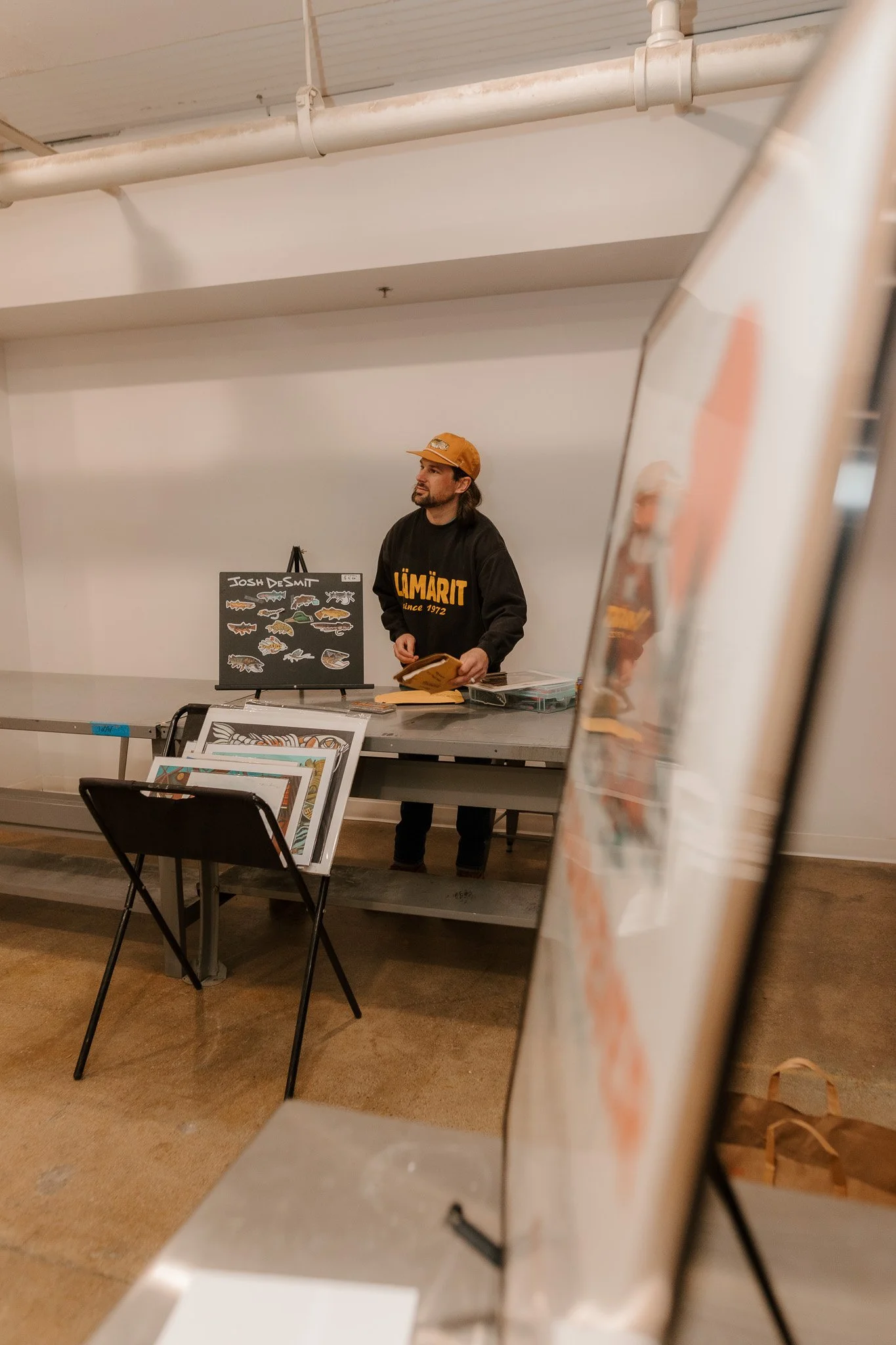 Man standing behind a table at a market stall, wearing a black sweatshirt with orange lettering and an orange cap, with various artwork and a blackboard display featuring fish illustrations in front of a white wall.